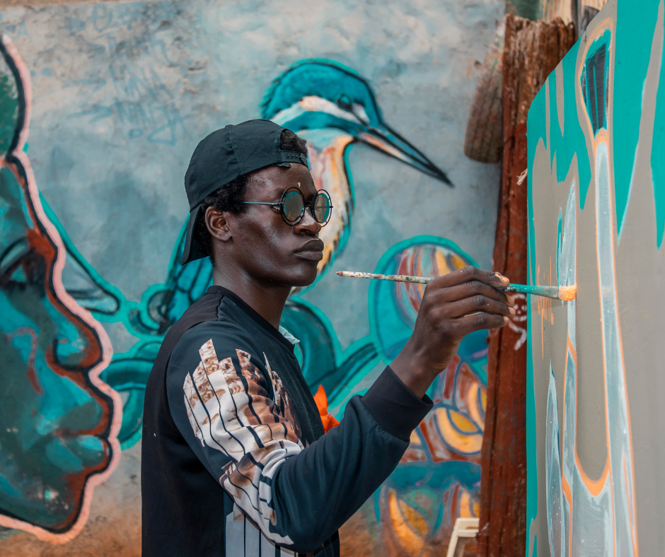 A young man wearing glasses, a black cap, and a black jacket with graphics, painting on a large canvas with a thin brush. The background features a mural of a heron and other abstract colorful designs.
