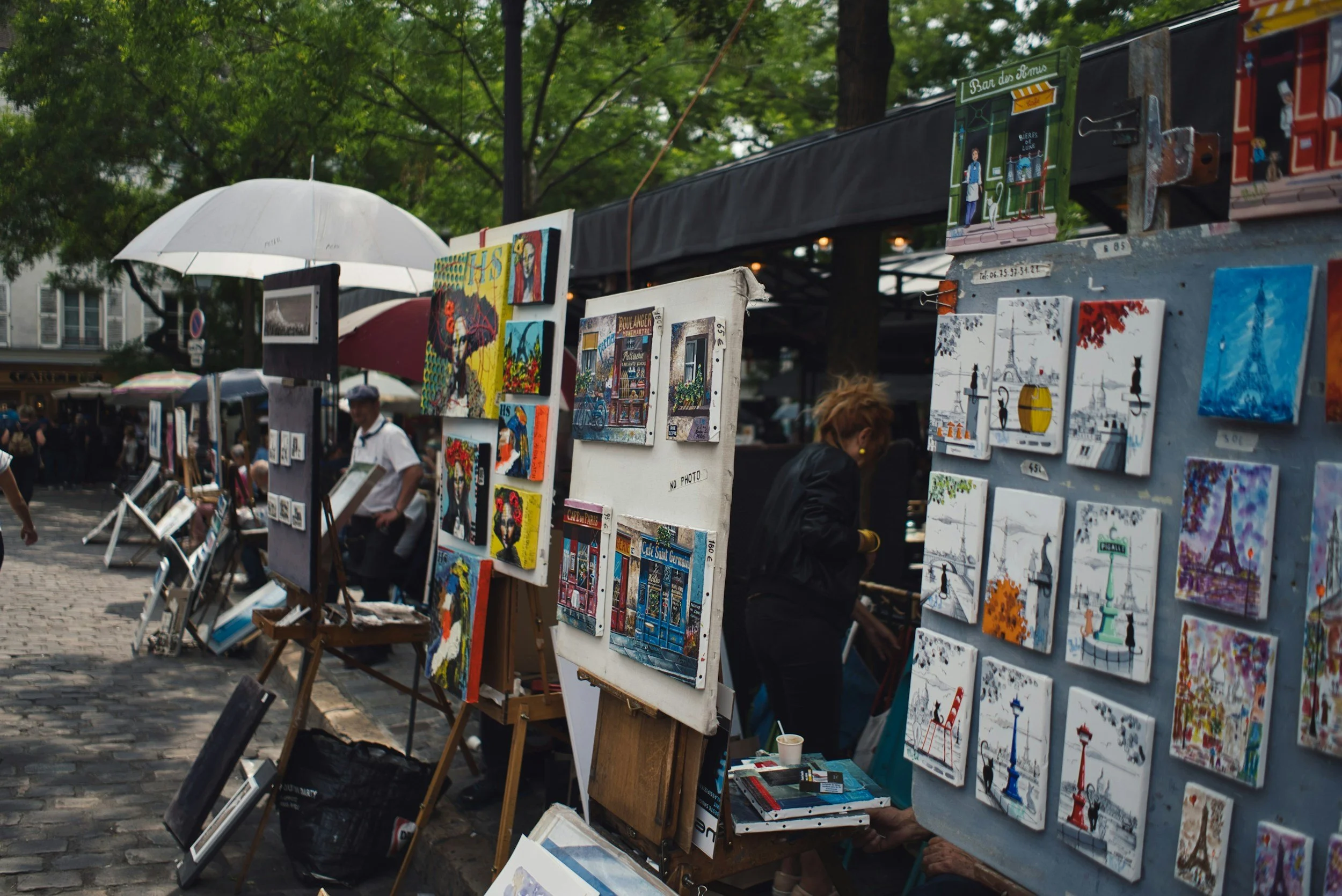 Outdoor art market with paintings on display, including Paris-themed scenes, with people browsing and umbrellas providing shade.