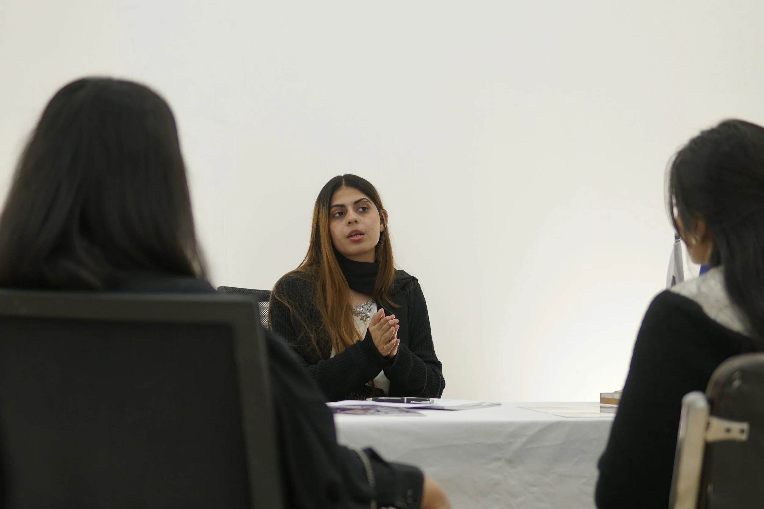 A woman is sitting at a table in front of two other women, speaking and gesturing with her hands during a discussion or meeting in a room with plain white walls.