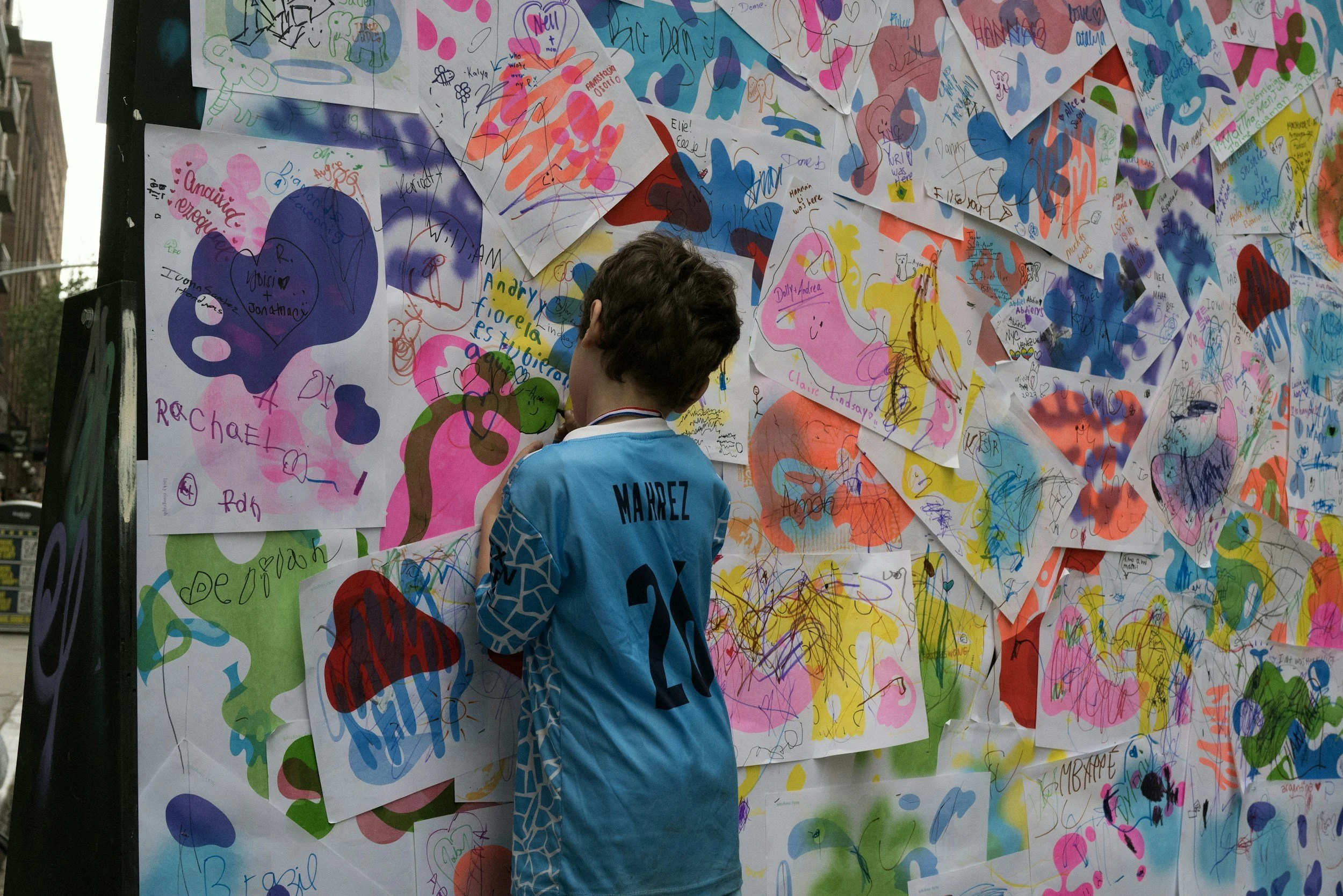A young person with curly brown hair, wearing a blue sports jersey with the name 'MARQUEZ' and number 21 on the back, stands in front of a large wall covered with colorful drawings and writings on white paper. The papers have various doodles, names, and drawings in bright colors like pink, purple, yellow, and blue, creating a vibrant collage.