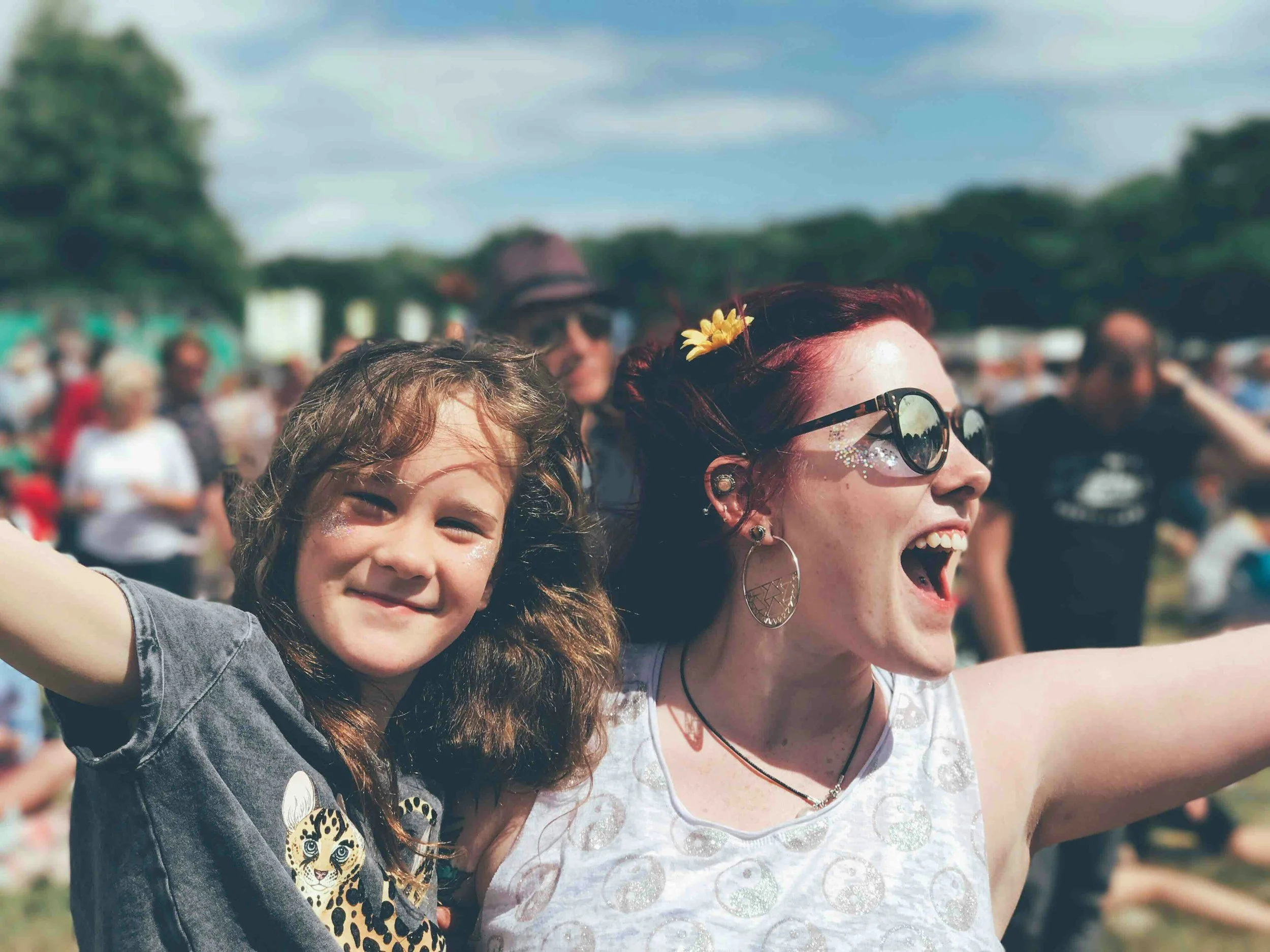 A young girl and a woman at an outdoor festival, smiling and enjoying the sunny day, with crowd and trees in the background.