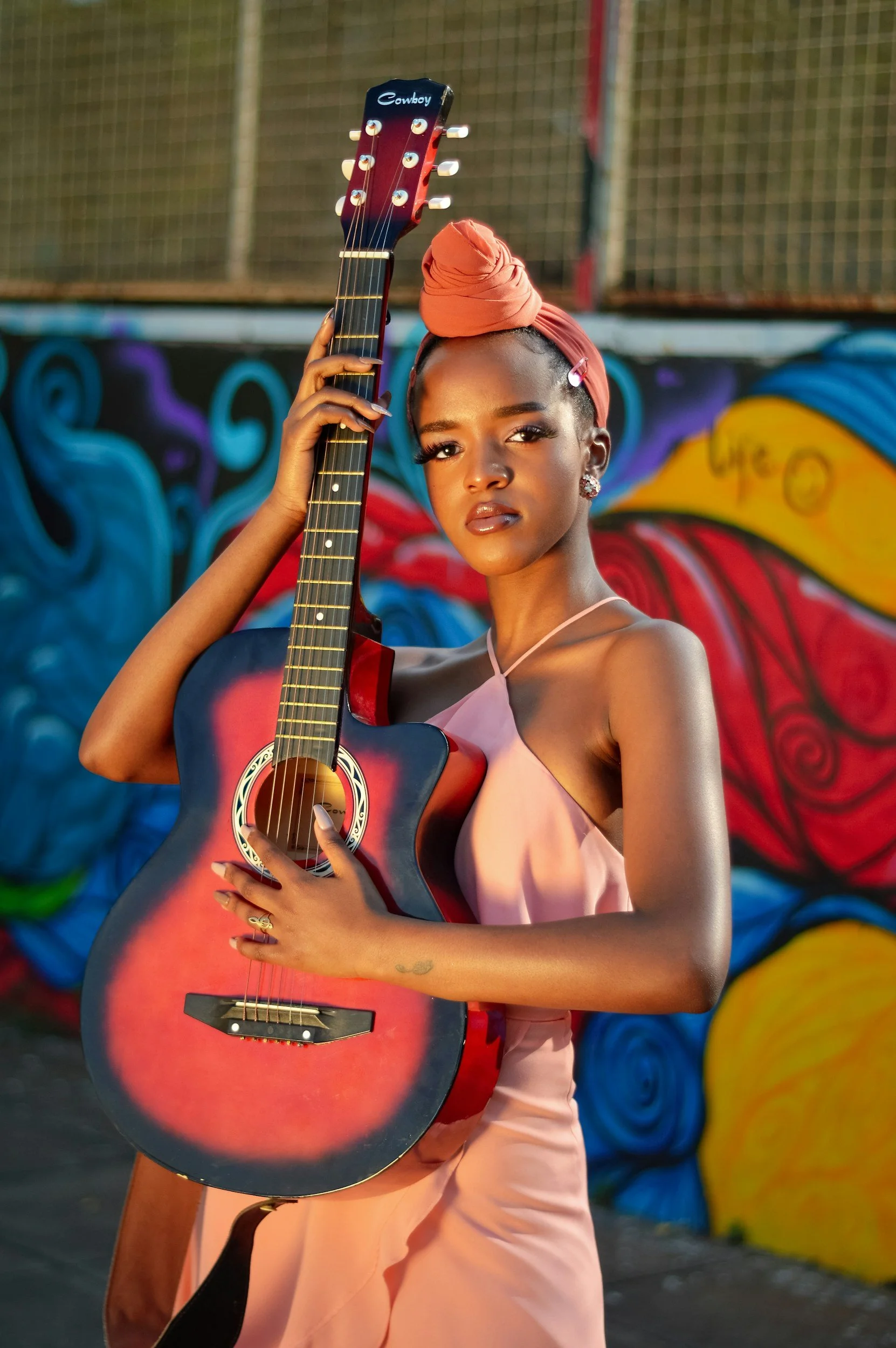 Young woman in pink dress with head wrap holding a red and black acoustic guitar, standing in front of colorful street art mural.