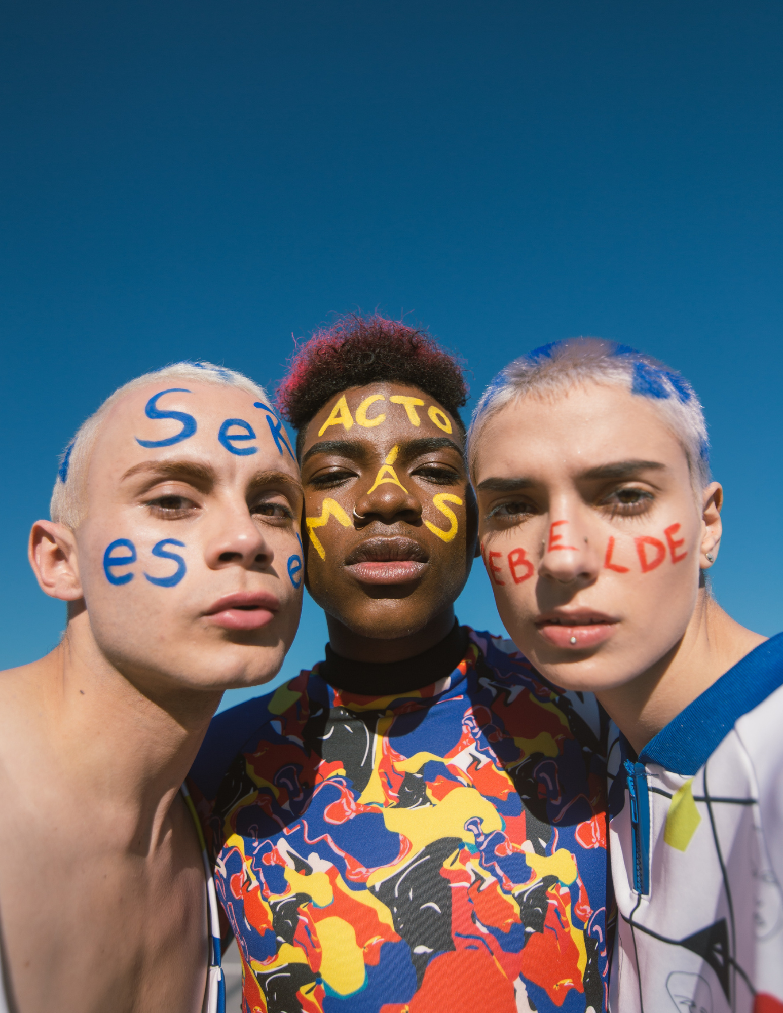 Three individuals with painted words and designs on their faces, standing close together against a clear blue sky.