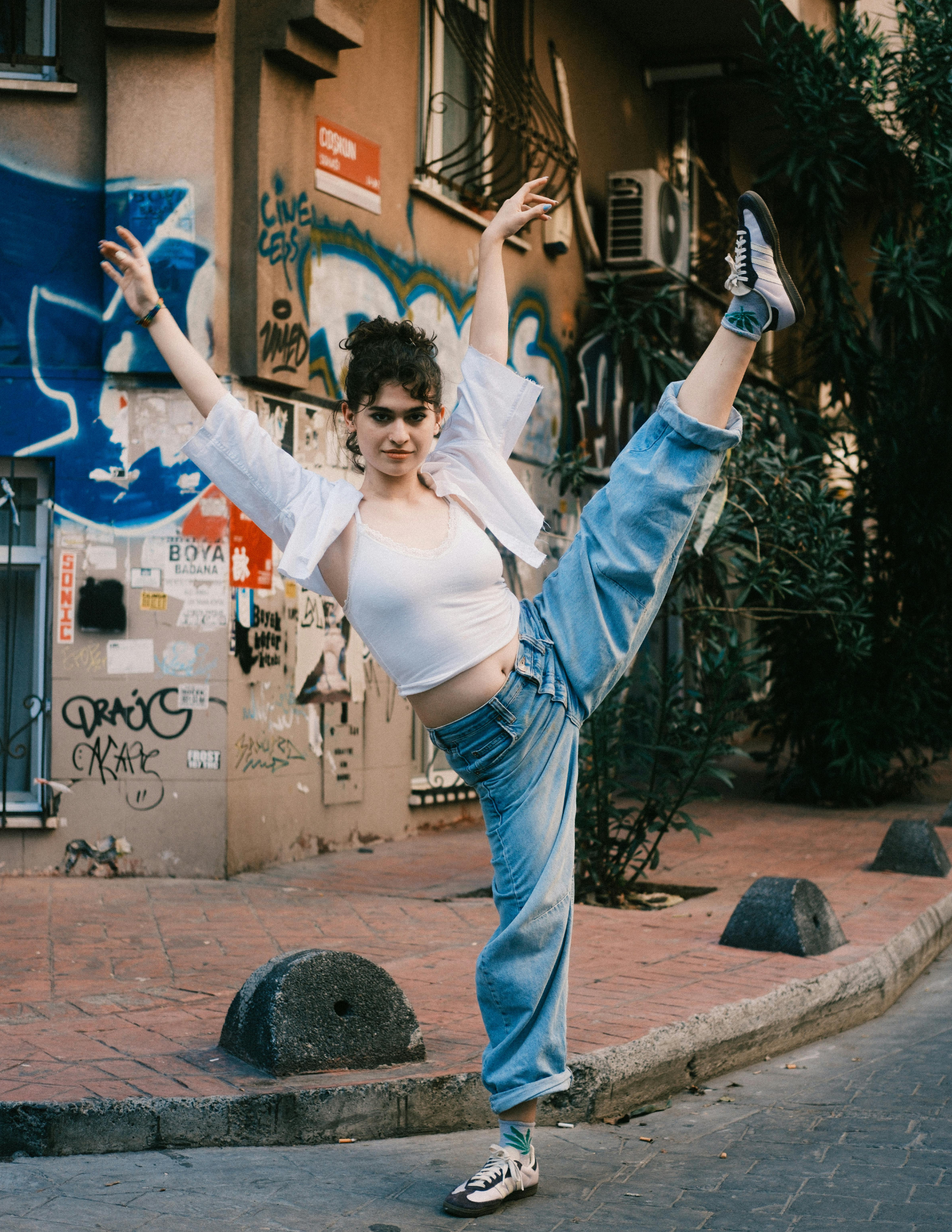 Young woman dancing or stretching on a city sidewalk with graffiti-covered walls behind her, wearing a white crop top, light blue jeans, and sneakers.