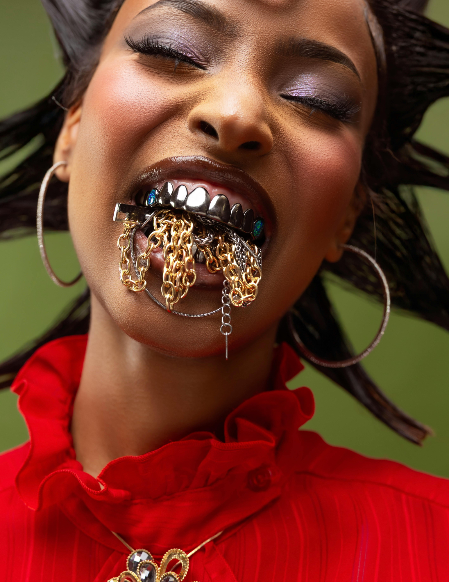 A woman with closed eyes, wearing makeup and large hoop earrings. She has a large lip ring with gold and silver chains dangling from her mouth, holding the chains in her teeth. She wears a red ruffled blouse and a gold necklace with black and white stones. Her hair is styled in loose waves.