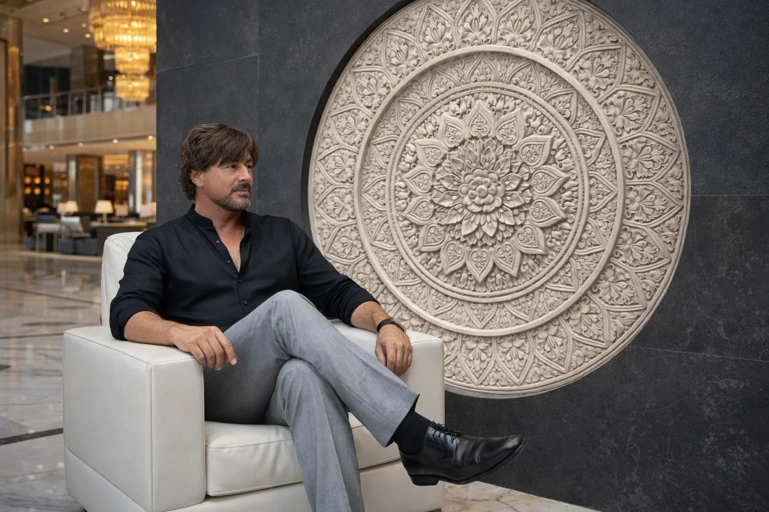 A man with brown hair, beard, and mustache sitting on a white armchair in a hotel lobby. He wears a black shirt, gray pants, and black shoes. Behind him is an intricate circular carved wall decoration with floral patterns.