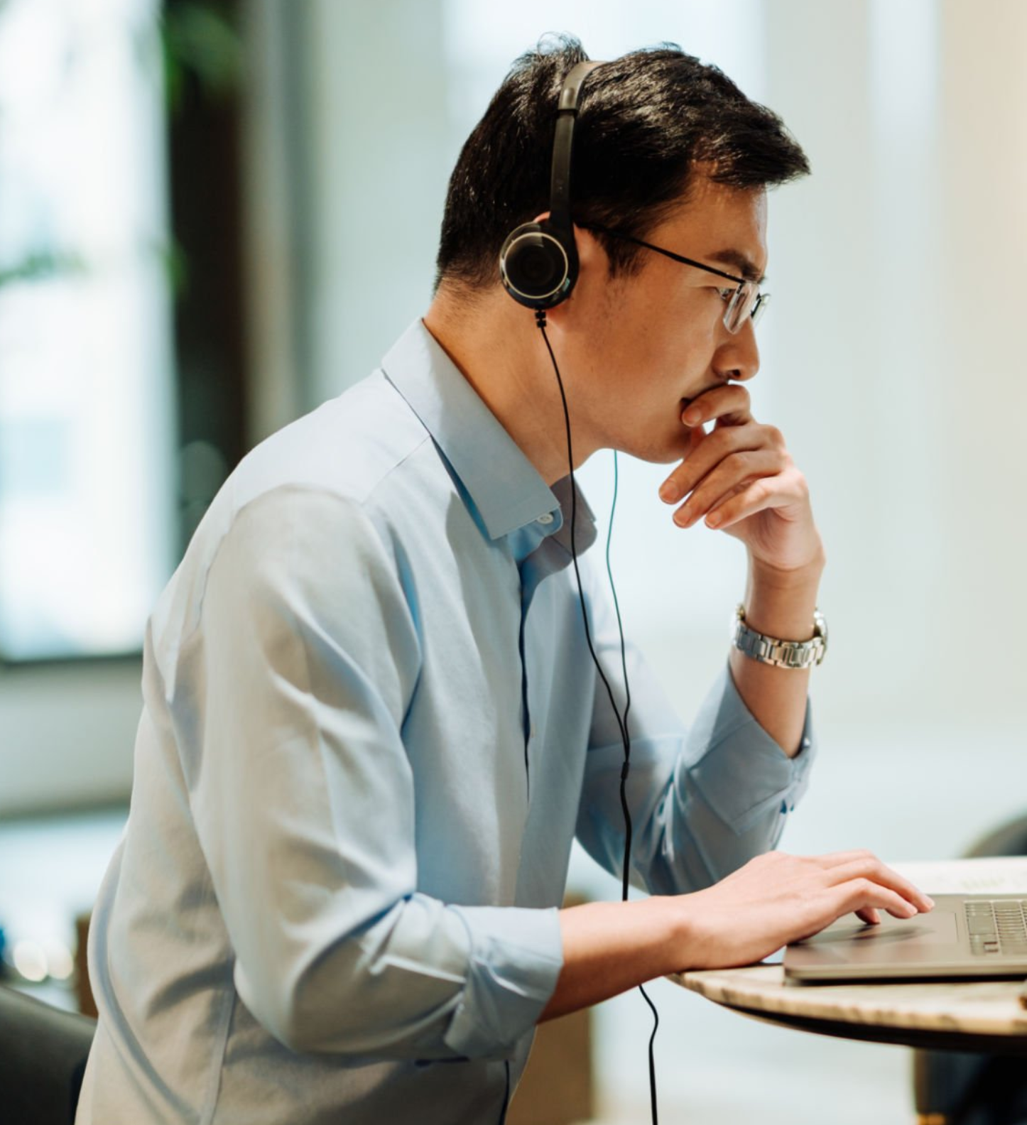眼鏡と腕時計を身につけた男性が、袖をまくった薄青色のシャツを着て机に座り、ヘッドホンを着用しながらノートパソコンを熱心に見ている。