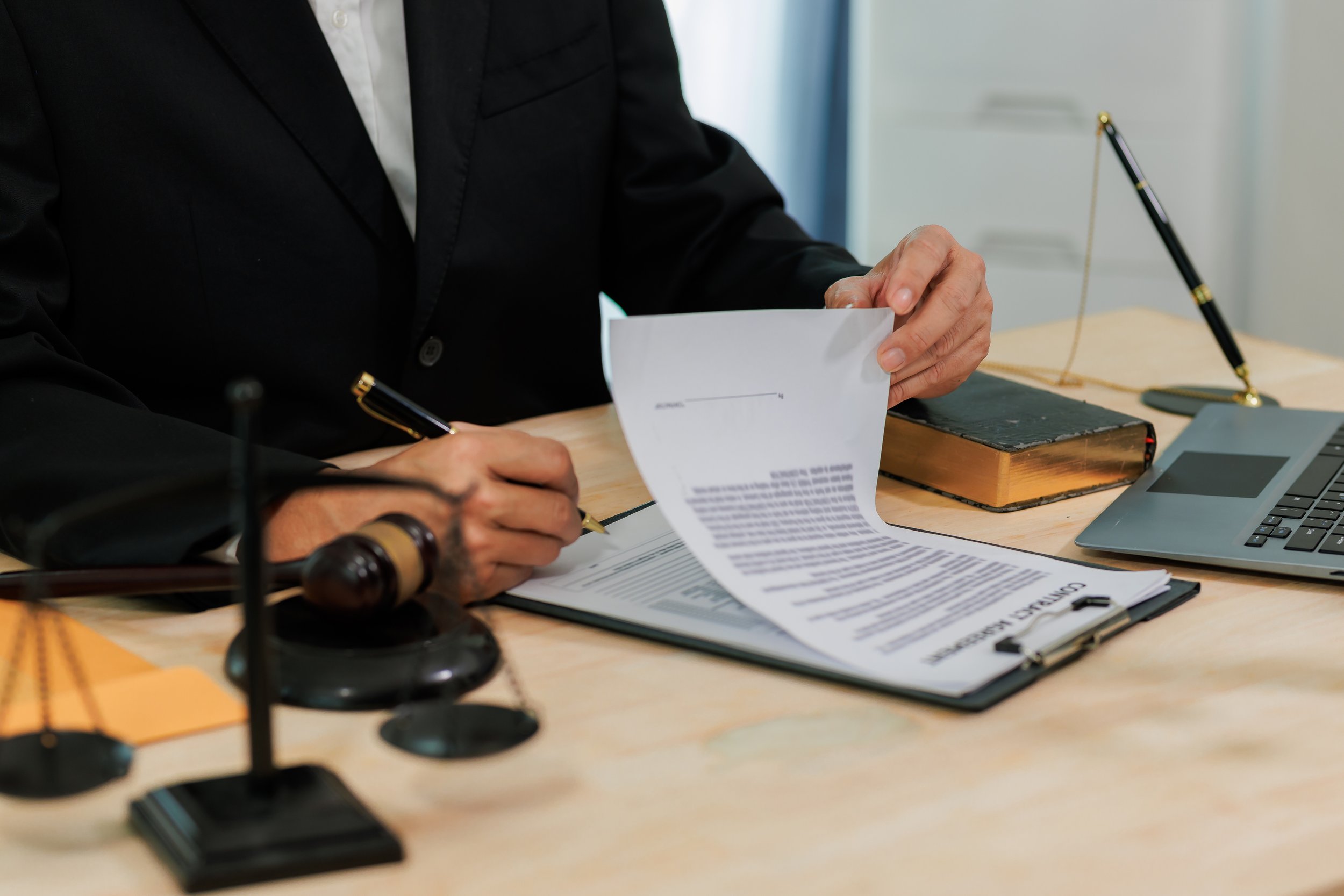 スーツ姿の人物が机で法的書類を確認している。机の上にはガベル、ノートパソコン、天秤が置かれている。