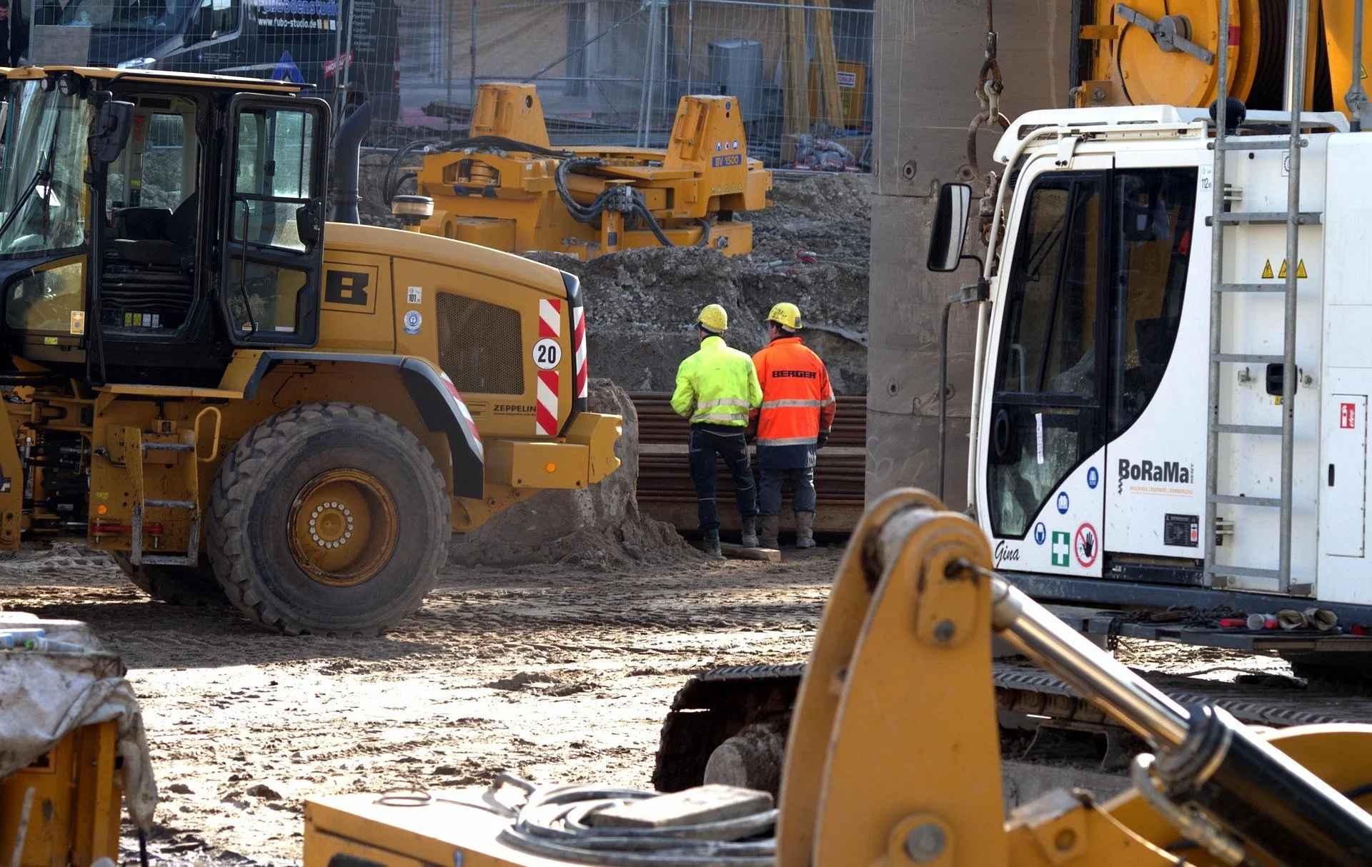 Trabajadores de construcción con chalecos y cascos de seguridad en obra, rodeados de maquinaria pesada, ejecutando labores de ingeniería y construcción.