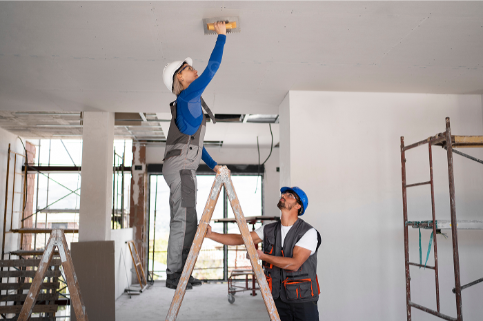 Two construction workers installing a ceiling light fixture in a building under construction, one standing on a ladder and the other holding the ladder.