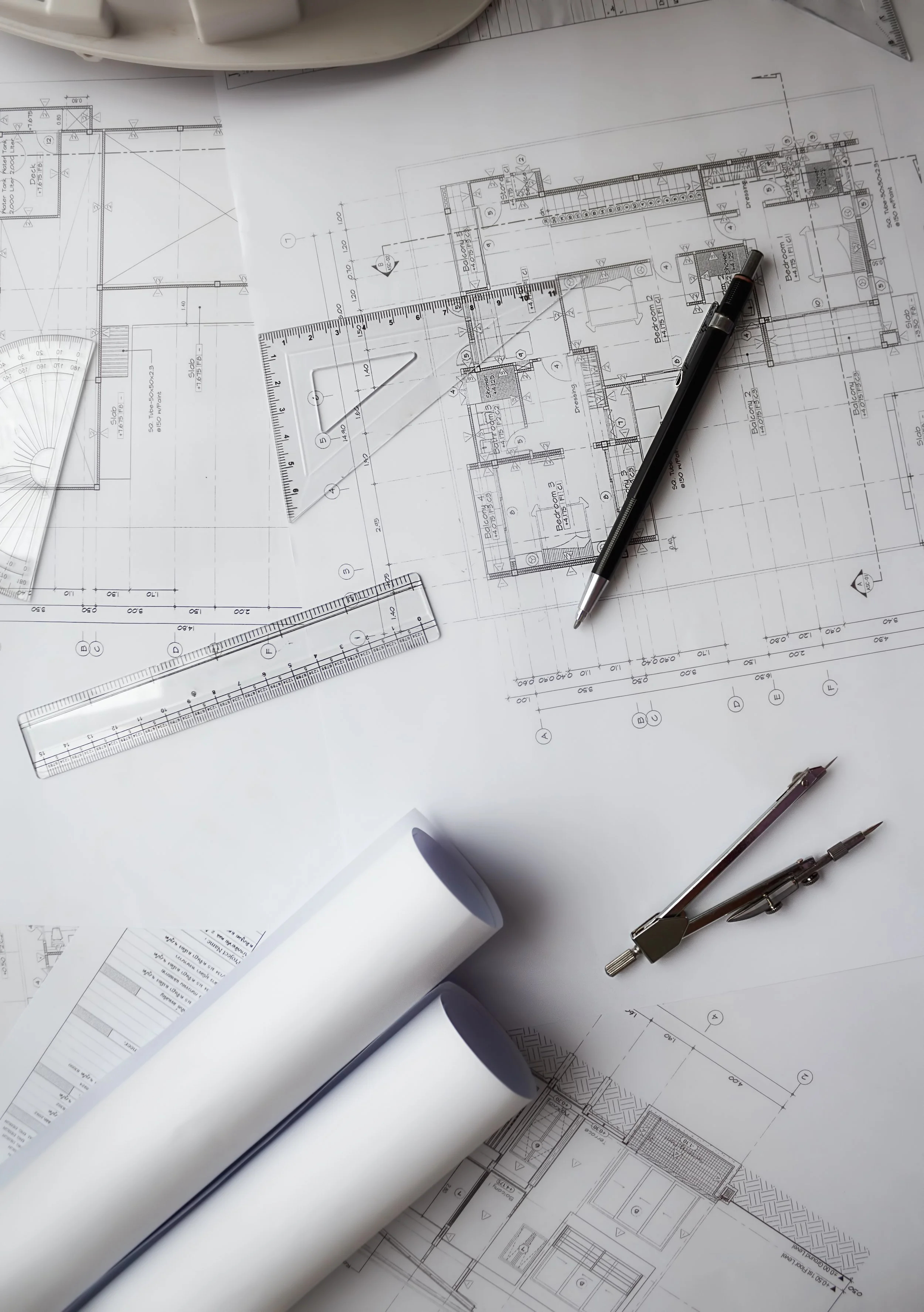 Architectural blueprints and drafting tools on a table, including rulers, compasses, a pen, and rolled-up plans.
