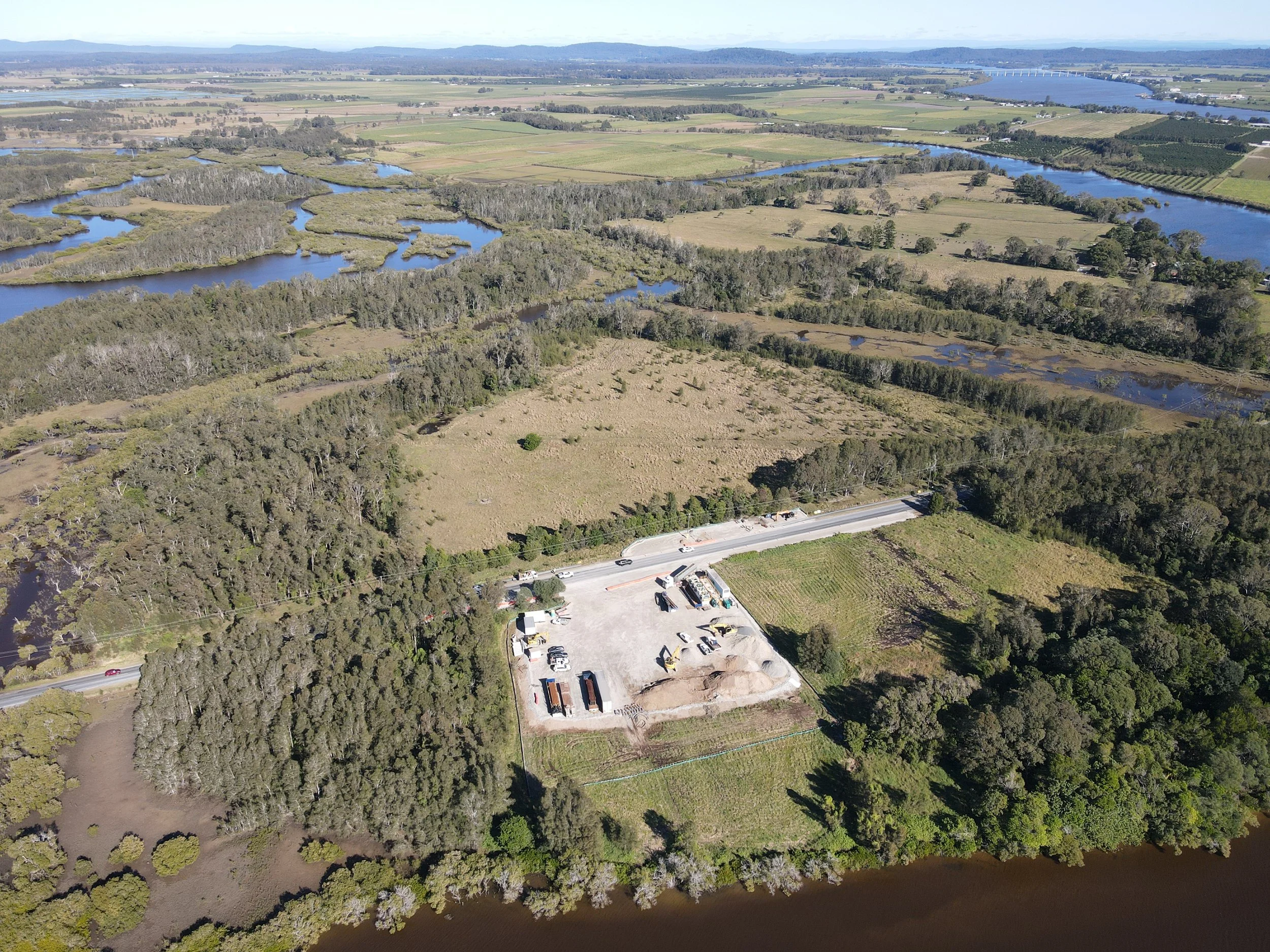 Aerial view of a landscape with a winding river, patches of wooded land, open fields, and a small industrial area with trucks and construction equipment at the bottom of the image.