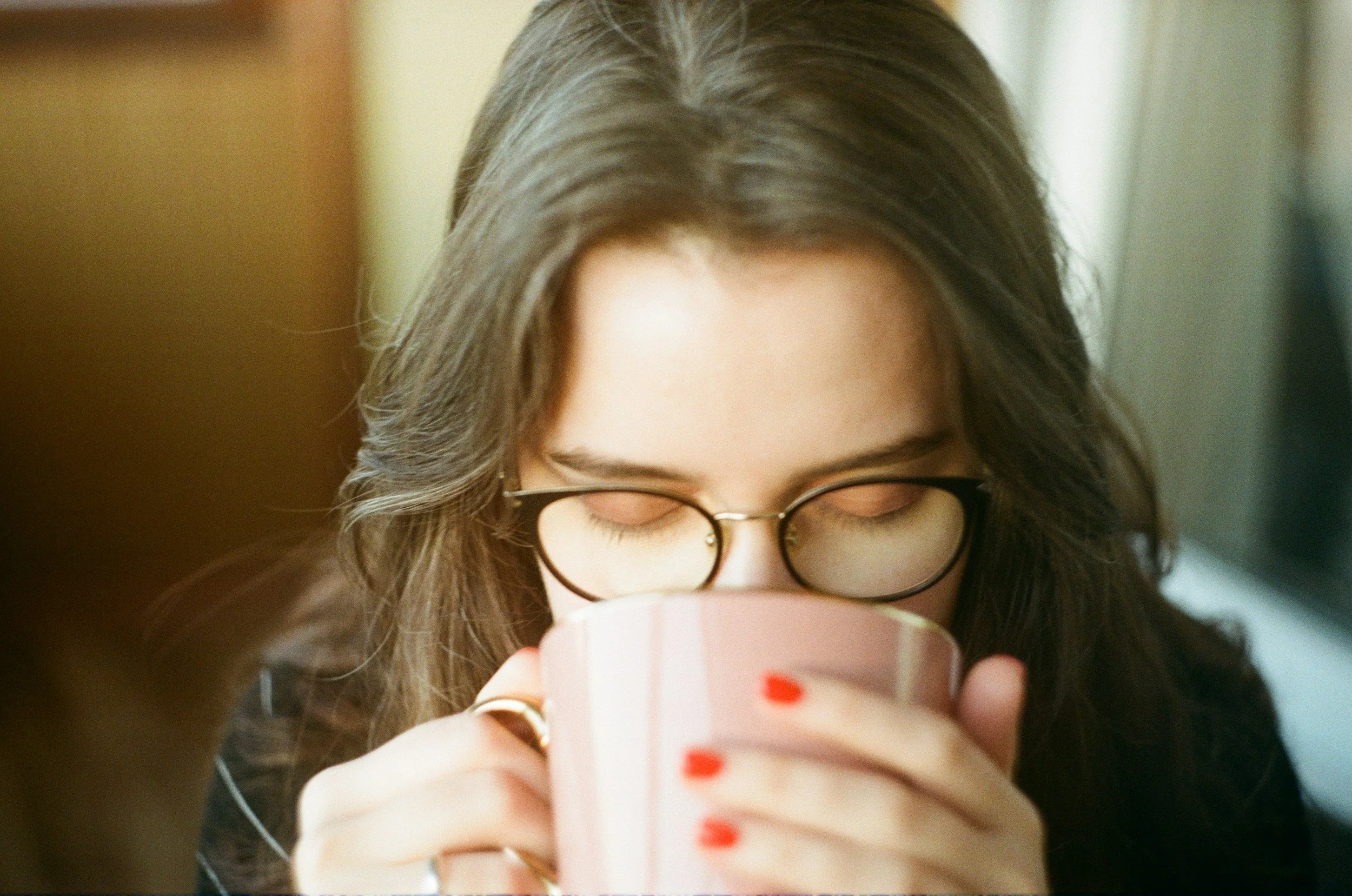 A young woman with long brown hair and glasses, wearing a dark shirt, drinking from a pink mug with red nails, in a cozy indoor setting.