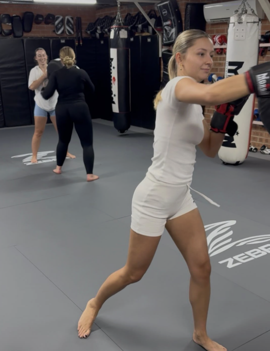 A woman practicing boxing in a gym, wearing white shorts and a white t-shirt, throwing a punch with boxing gloves on, with two other women boxing in the background.