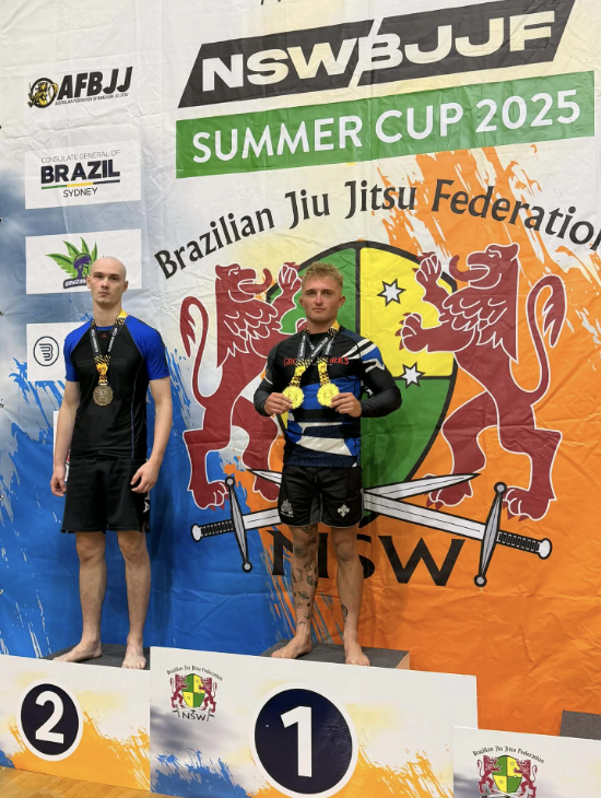 Two male athletes standing on a winners' podium at a Brazilian Jiu Jitsu competition, with medals around their necks. The athlete in the center on the first-place platform holds a gold medal, while the athlete on the second-place platform has a silve