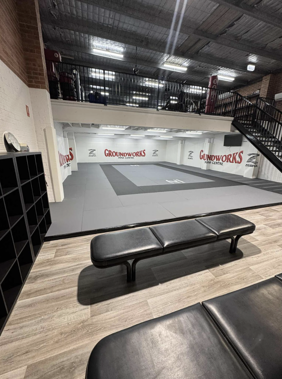 An indoor martial arts gym with a tatami mat area, benches, and shelving. The walls are decorated with 'Groundworks MMA Centre' and 'Zebra' logos.