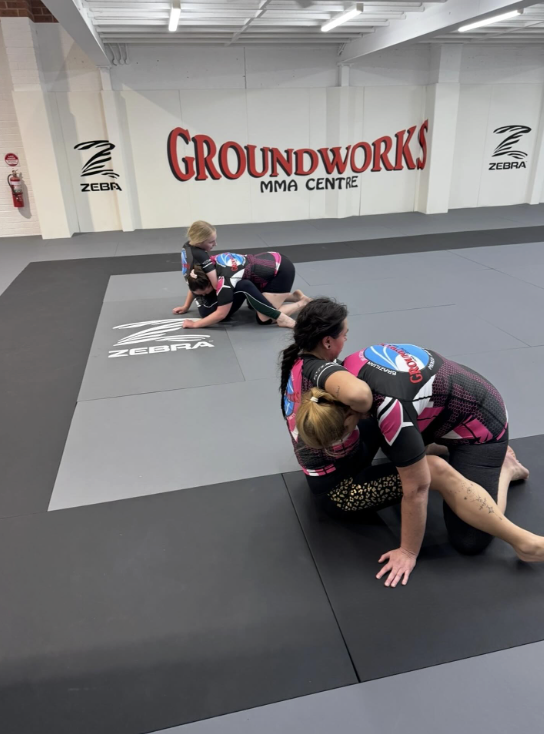 Two women practicing Brazilian Jiu-Jitsu on mats in a gym with a sign that reads Groundworks MMA Centre