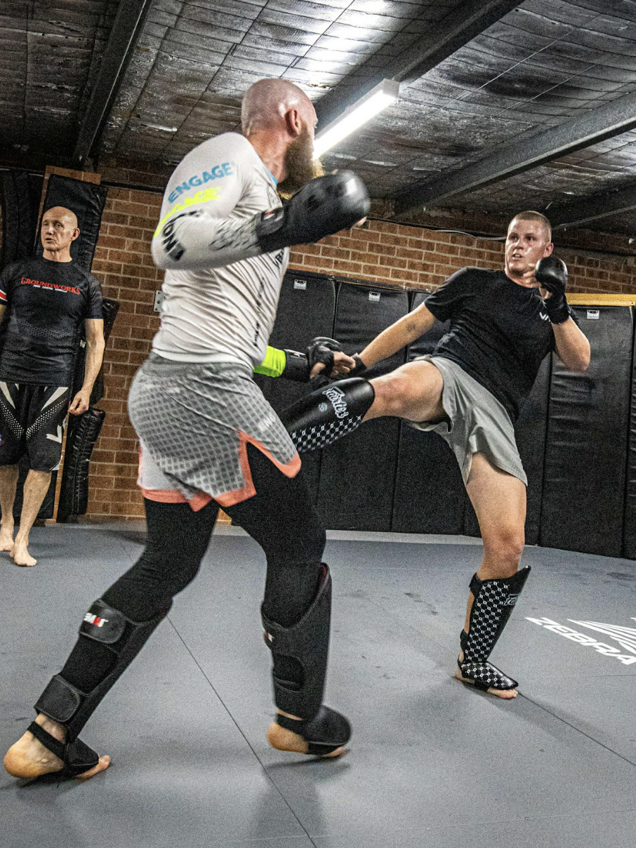 Two men practicing martial arts sparring with one executing a high kick on the other in a gym, with a third man observing.