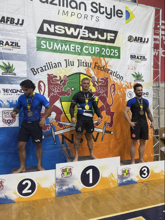 Three male athletes standing on a winners' podium with medals after a Brazilian Jiu Jitsu competition, with a large banner behind them that reads 'Amazon Style Imports NSWBJJ Summer Cup 2025' and features logos of the Brazilian Jiu Jitsu Federation, 