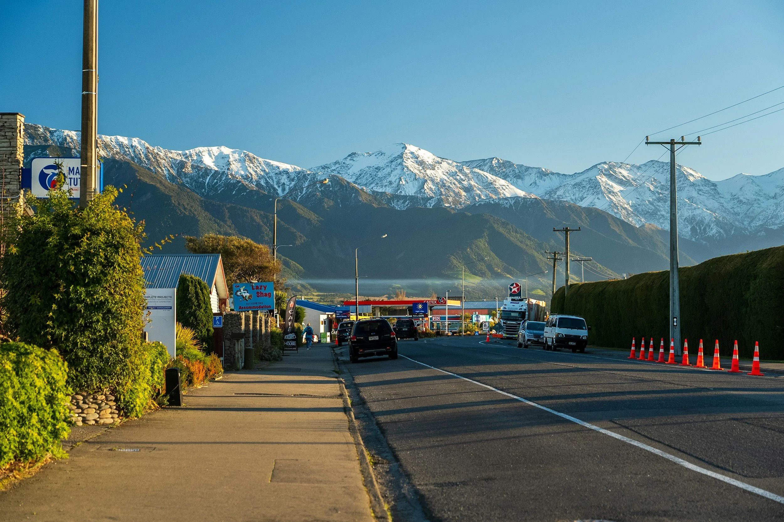 A small roadside town with a mountainous, snow-capped landscape in the background, featuring a gas station and parked cars on a clear day.