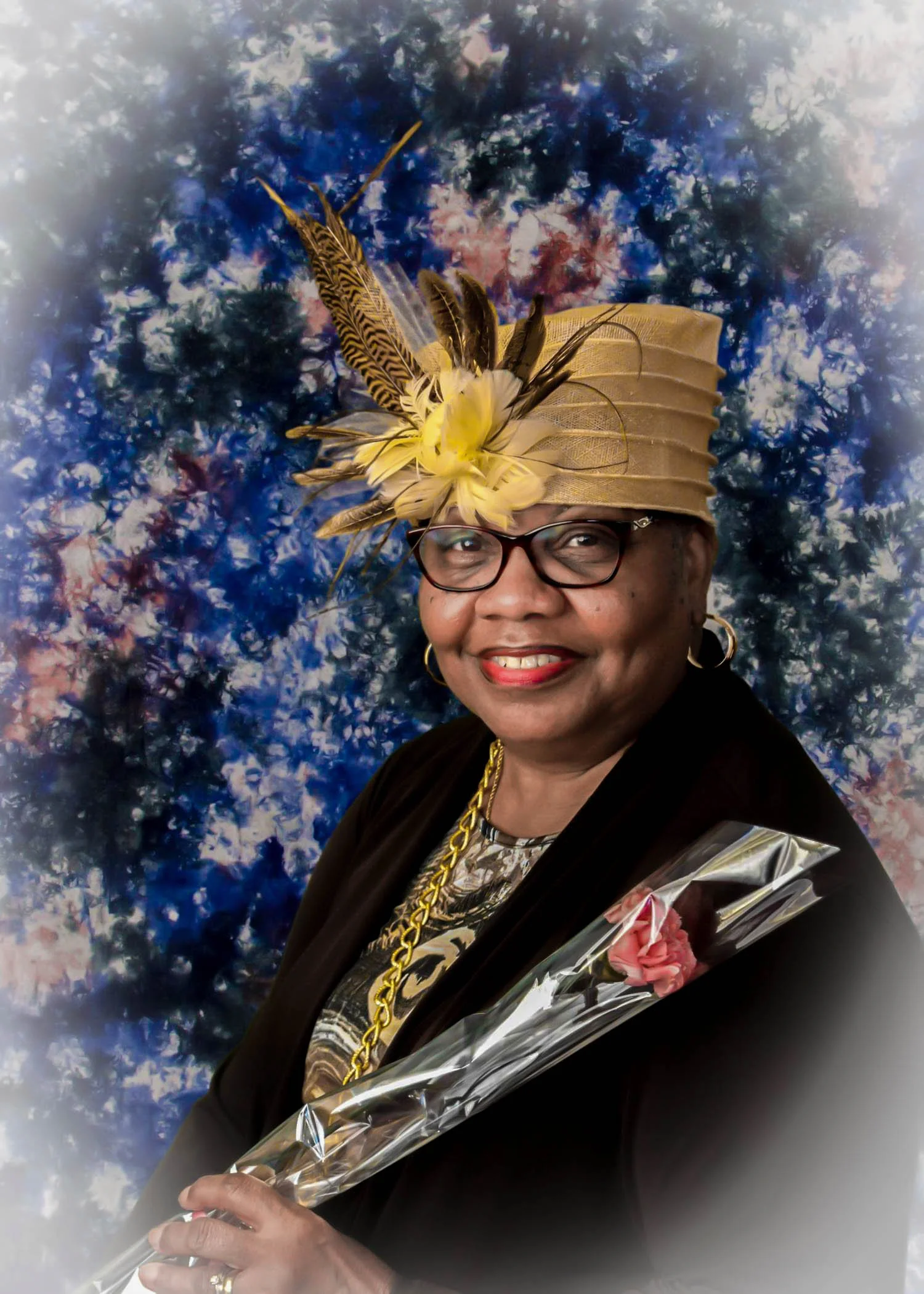 A smiling woman wearing glasses, a gold hat with feathers, black clothing, gold jewelry, holding a wrapped bouquet, against a blue and pink abstract background.