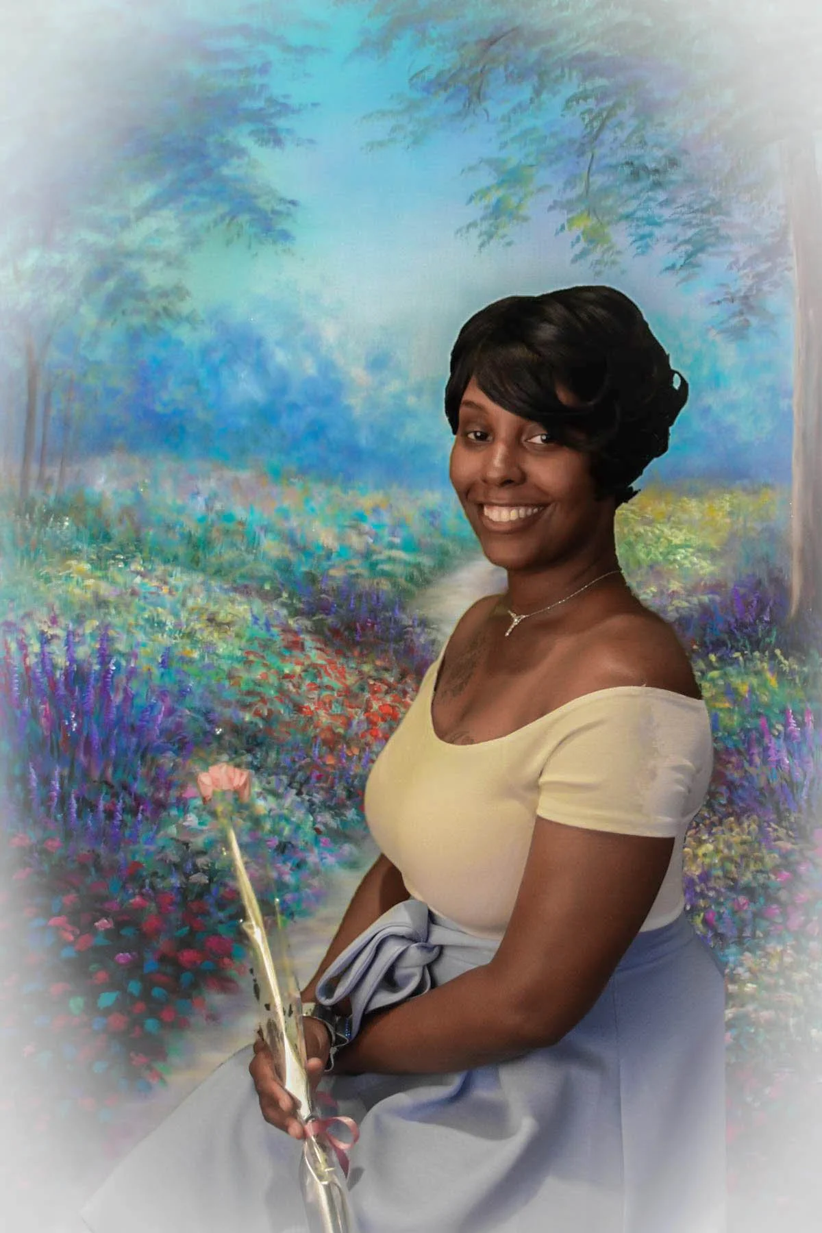 A smiling woman with short black hair and a yellow off-the-shoulder top holding a single pink flower, standing in front of a colorful, painted landscape background.