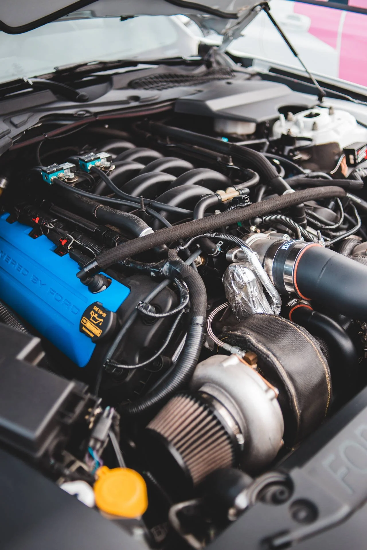Close-up of a high-performance car engine with various parts, hoses, and a blue valve cover marked 'Powered by Ford'.
