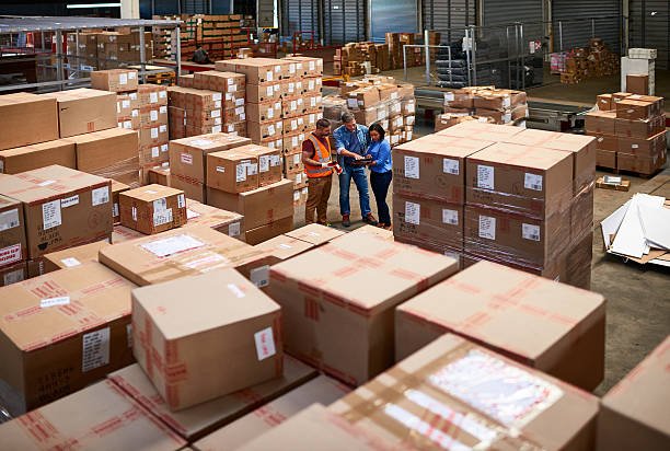 Warehouse with large stacks of cardboard boxes and three workers examining documents.