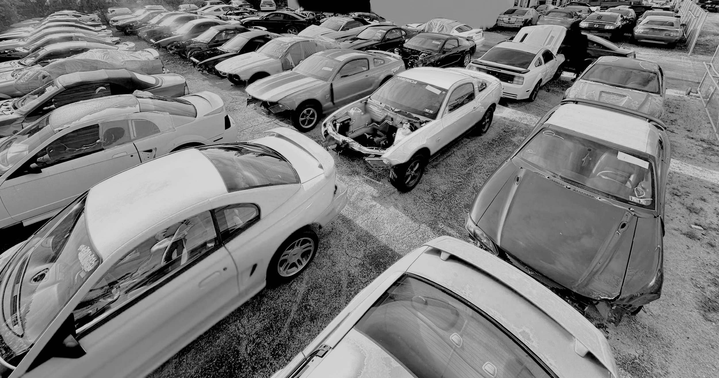 A black and white photo of a lot filled with various cars, including some damaged and some in good condition, parked closely together.