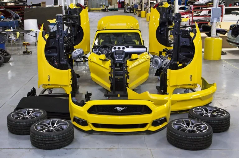 Disassembled yellow Ford Mustang sports car in a factory or workshop with four tires placed in front, showing the car's front, side, and interior components.