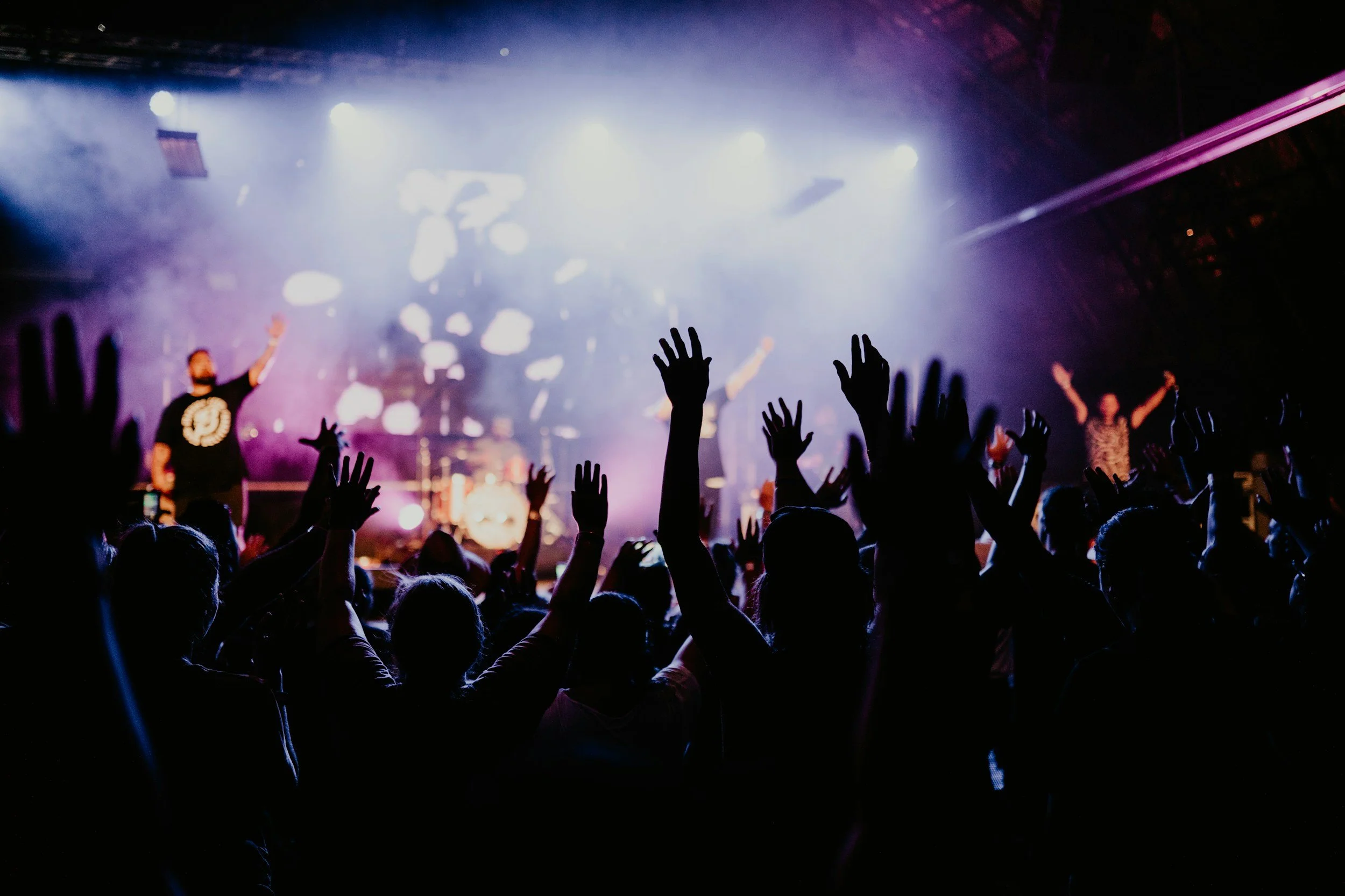Crowd at a live concert with hands raised, stage lighting, and performers in the background.