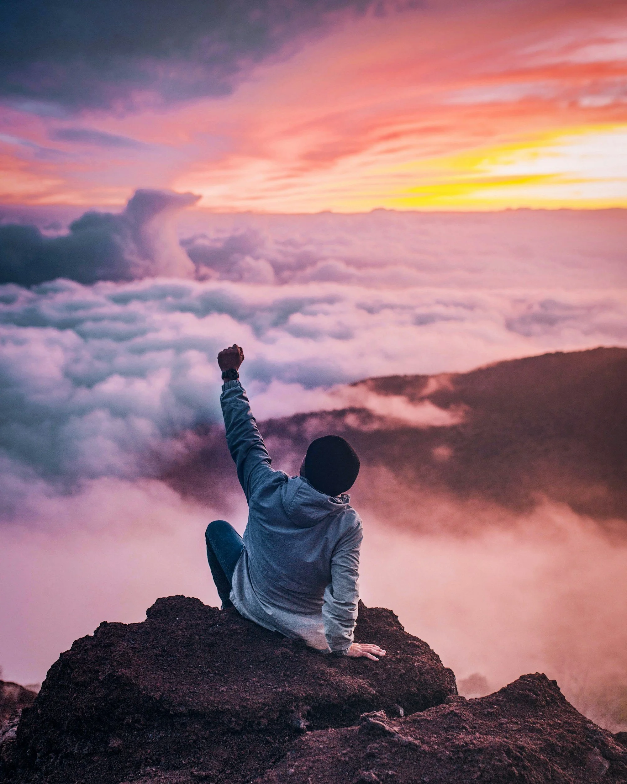 A person sitting on a rocky cliff at sunset above the clouds, raising one fist in the air, with colorful pink, purple, and yellow sky.