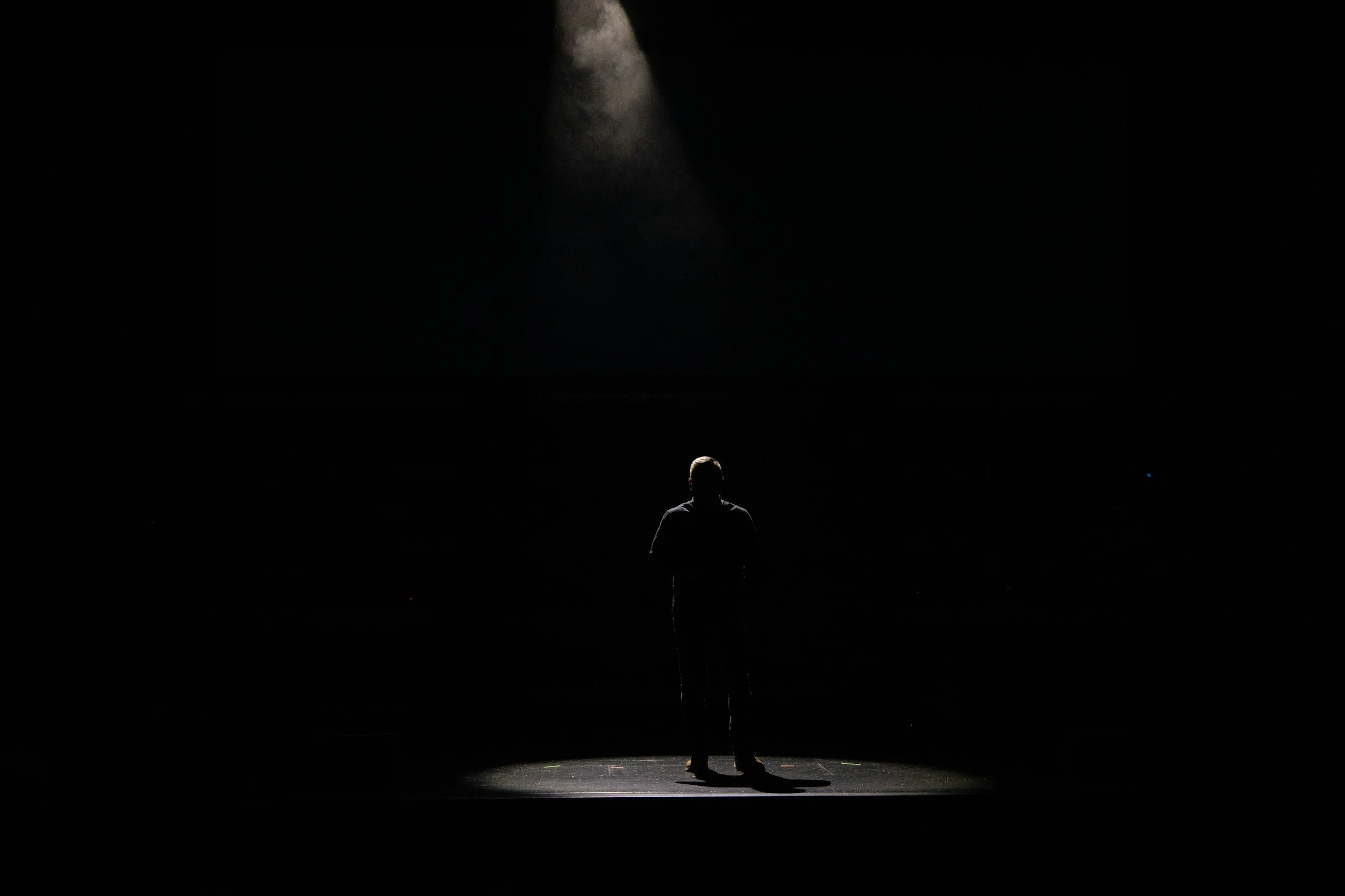 A person stands alone on a stage in a dark theater, illuminated by a single spotlight overhead.