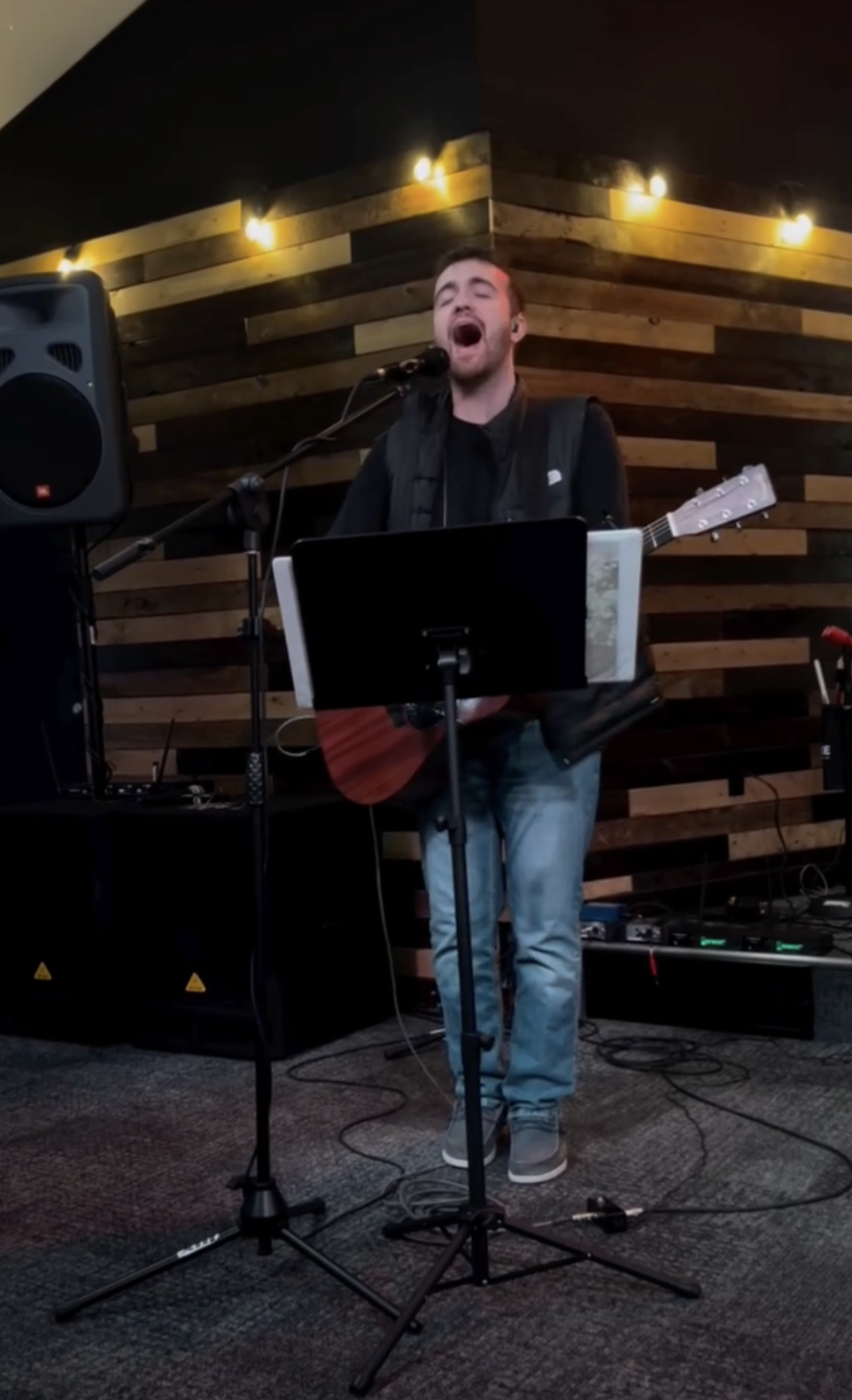 A man singing into a microphone while playing an acoustic guitar, standing in front of a wooden wall with warm lighting.