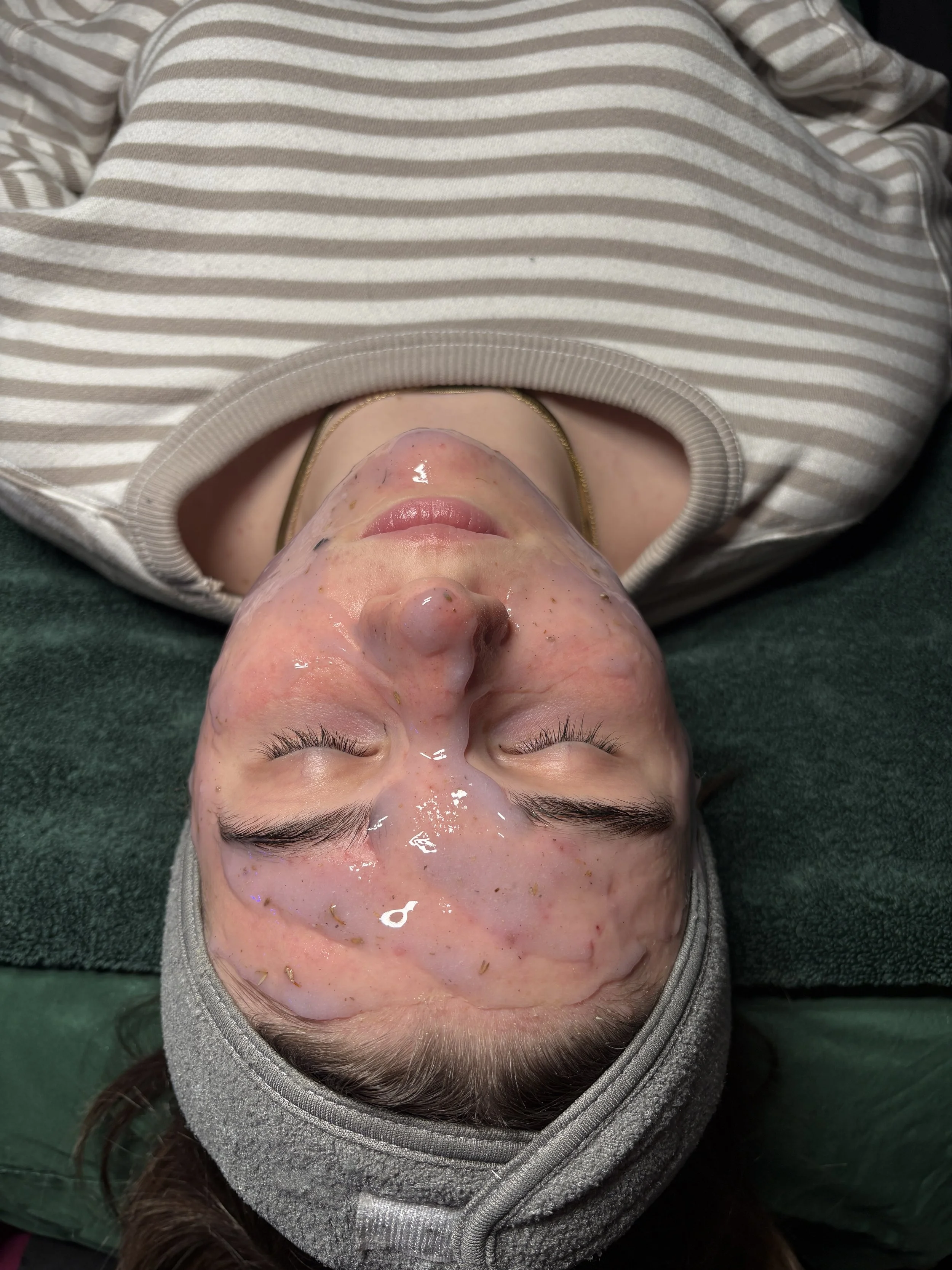 A woman lying down with her eyes closed, wearing a gray striped hoodie and a gray headband. Her face is covered with a translucent facial mask with small particles, and she appears to be in a spa or skincare treatment setting.