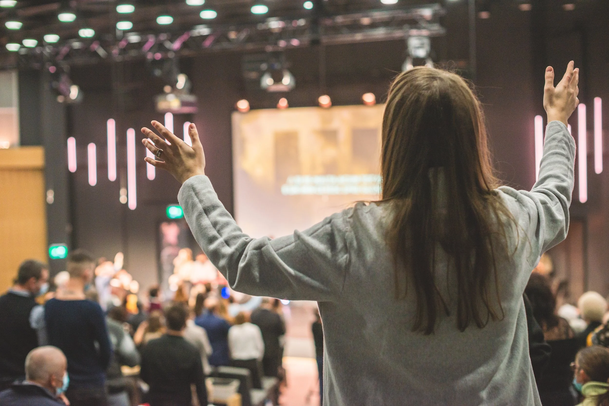 A woman with long brown hair and a gray sweatshirt is raising her hands in the air while facing a stage with a large screen and purple lights, in front of an audience at an indoor event or conference.