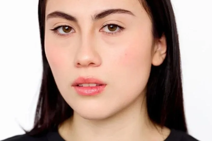 Close-up of a young woman with fair skin, dark brown hair, and hazel eyes looking directly at the camera, wearing a black top.