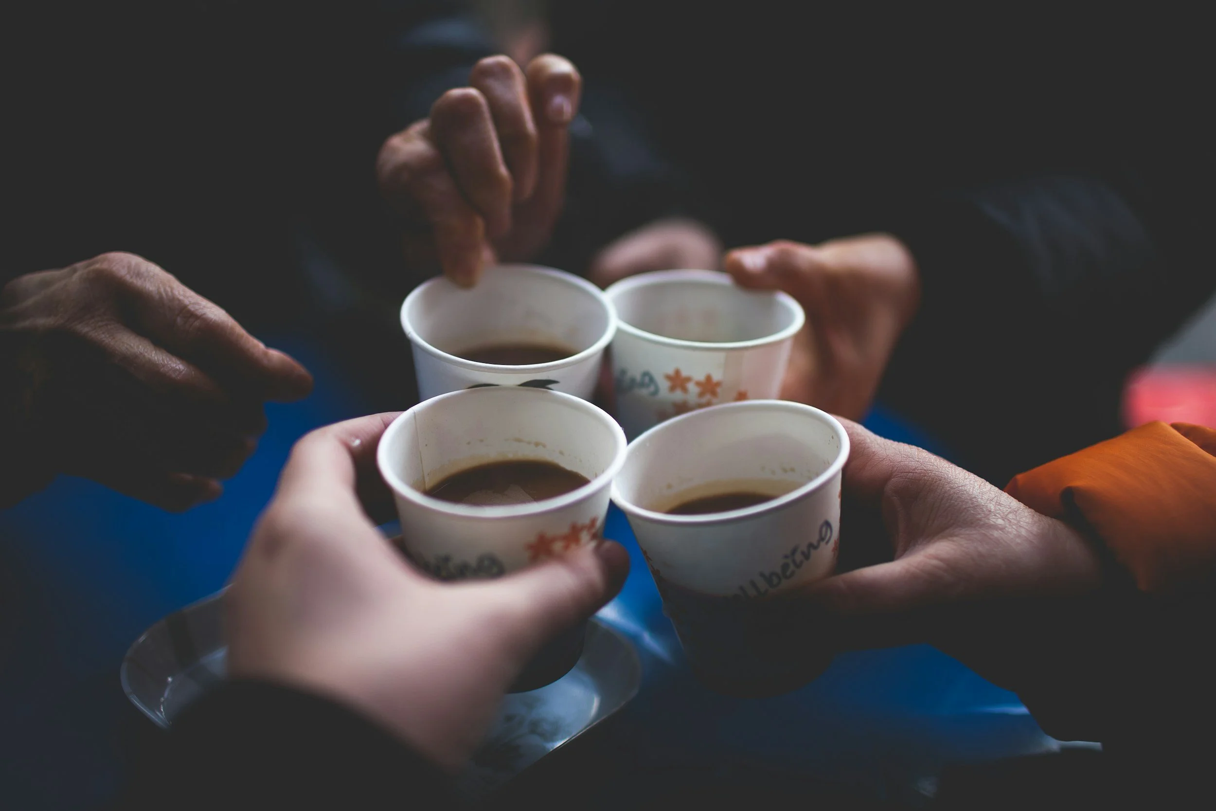 Four people holding small paper cups with coffee for a toast, close-up view of their hands.