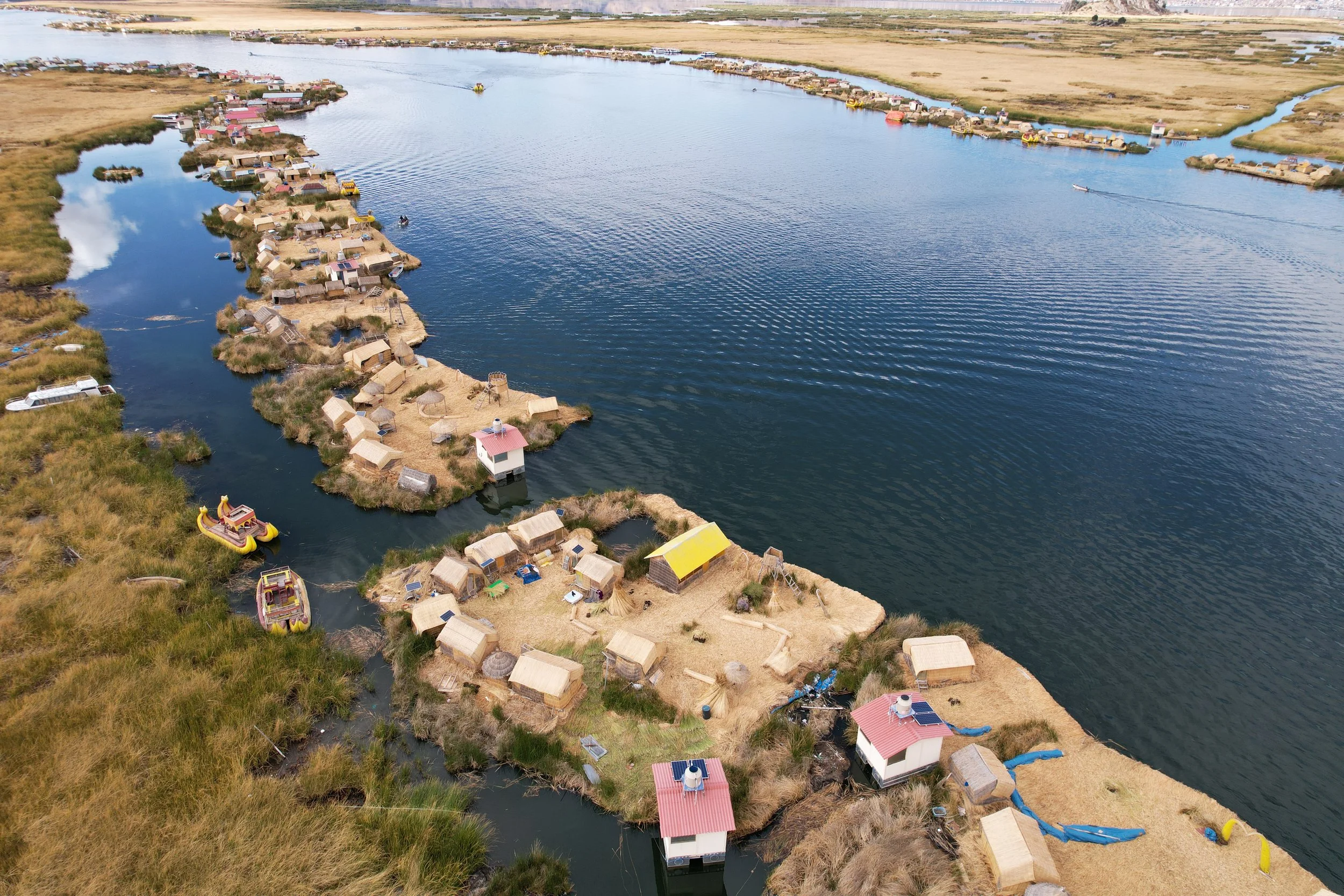 Aerial view of a small riverside village with houses and boats, surrounded by grasslands and water.
