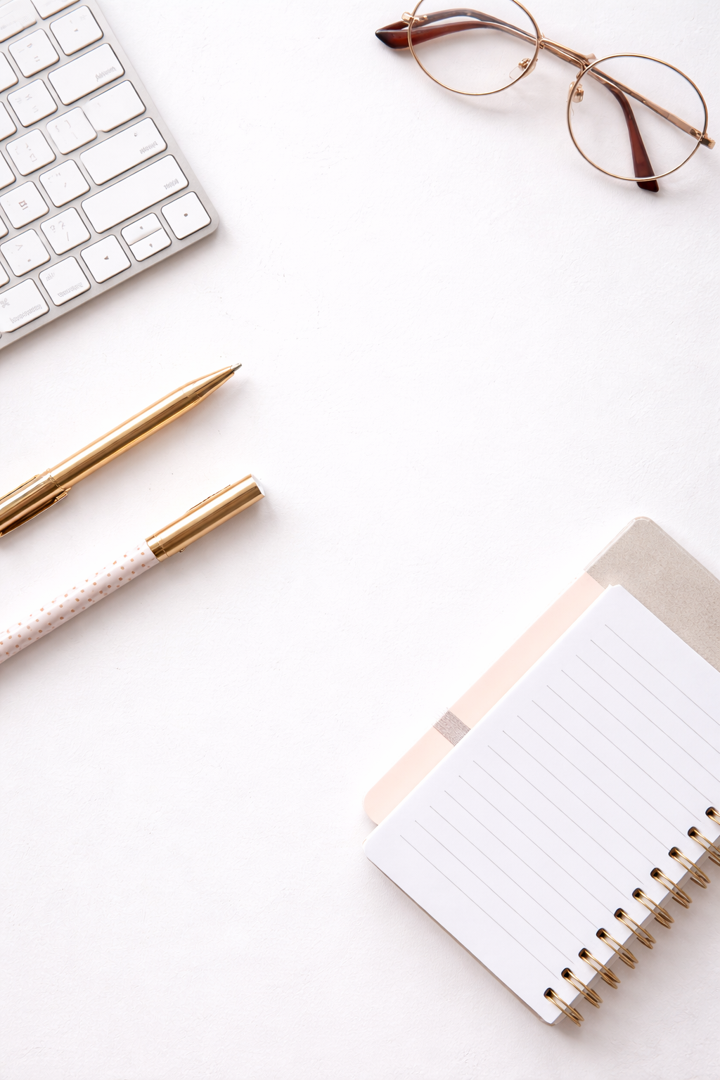 Minimalist vertical workspace with a keyboard, glasses, pens, and notebook on a white desk.