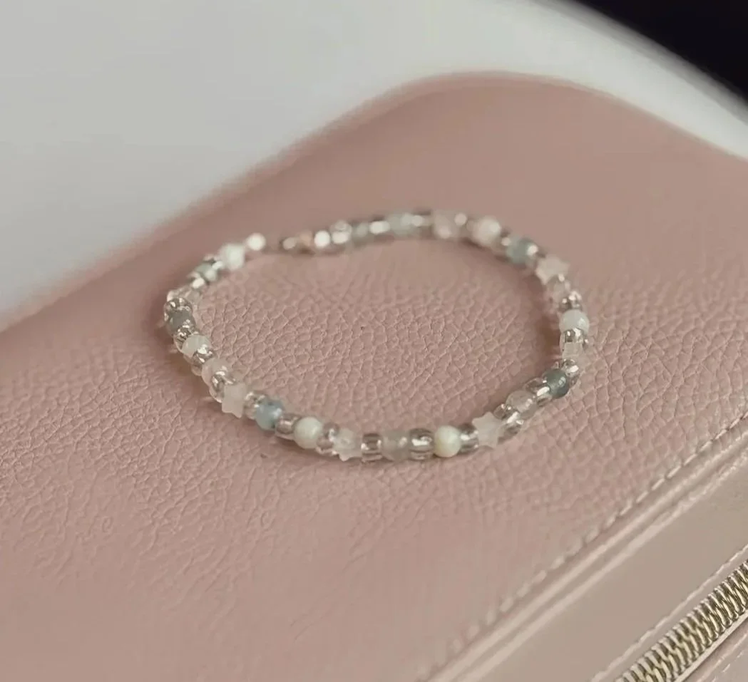 A delicate beaded bracelet with white, gray, and clear beads resting on a pink textured jewelry box.