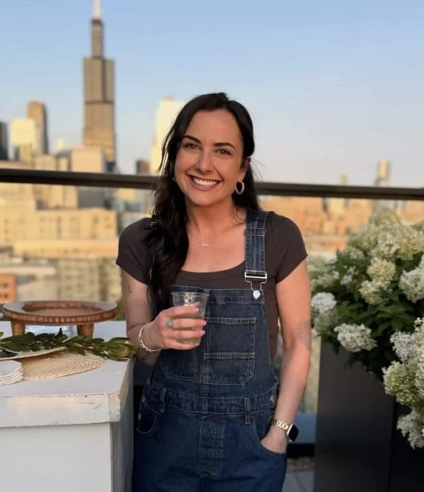 A woman with dark hair, wearing earrings and a bracelet, standing on a rooftop with a city skyline in the background. She is smiling, holding a drink, and dressed in a dark T-shirt and denim overalls. There are white flowers and a table with some decorative items nearby.