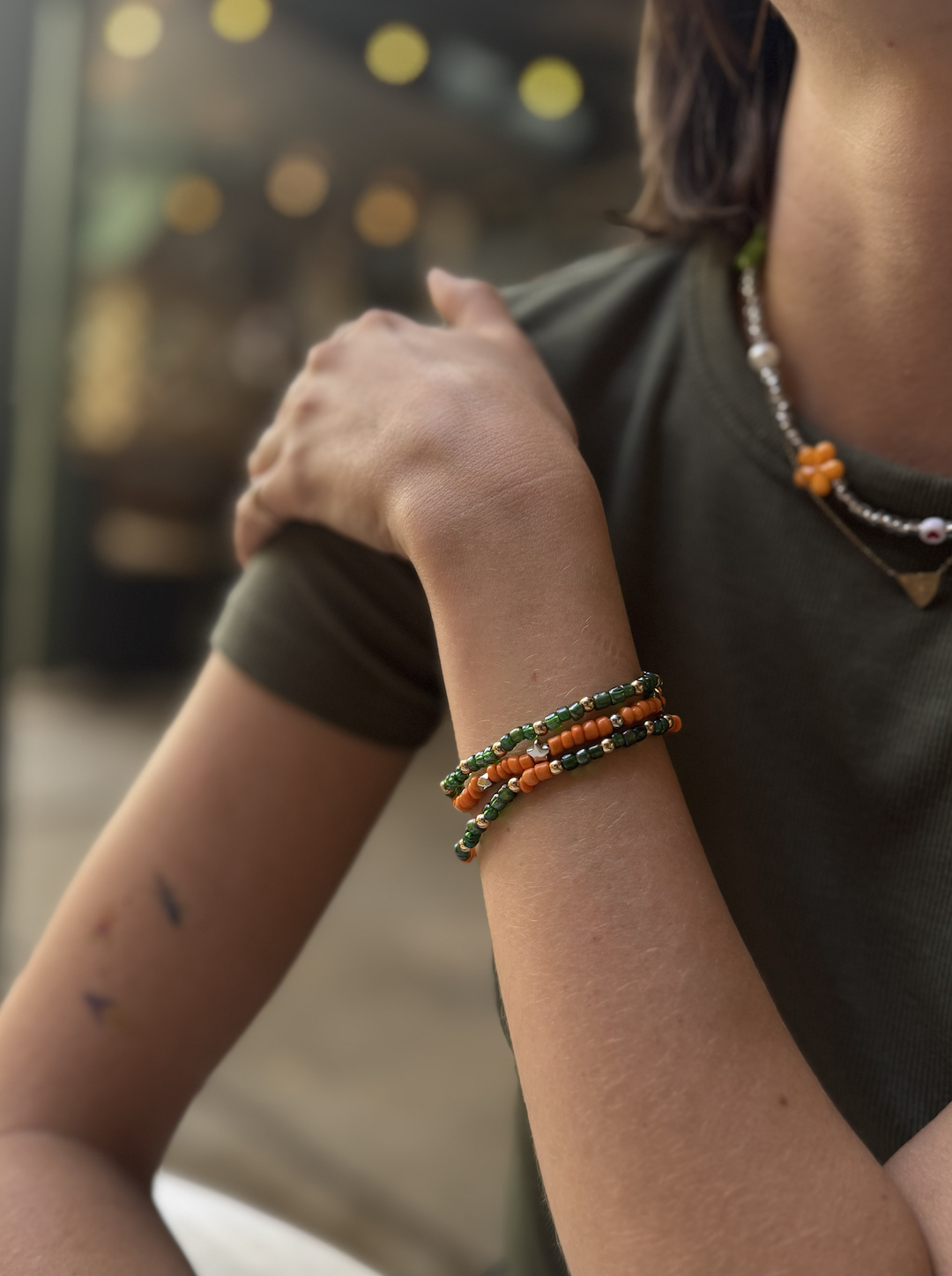 Close-up of a person's arm and shoulder, wearing beaded jewelry including bracelets and a necklace, with a blurred background of warm lights.