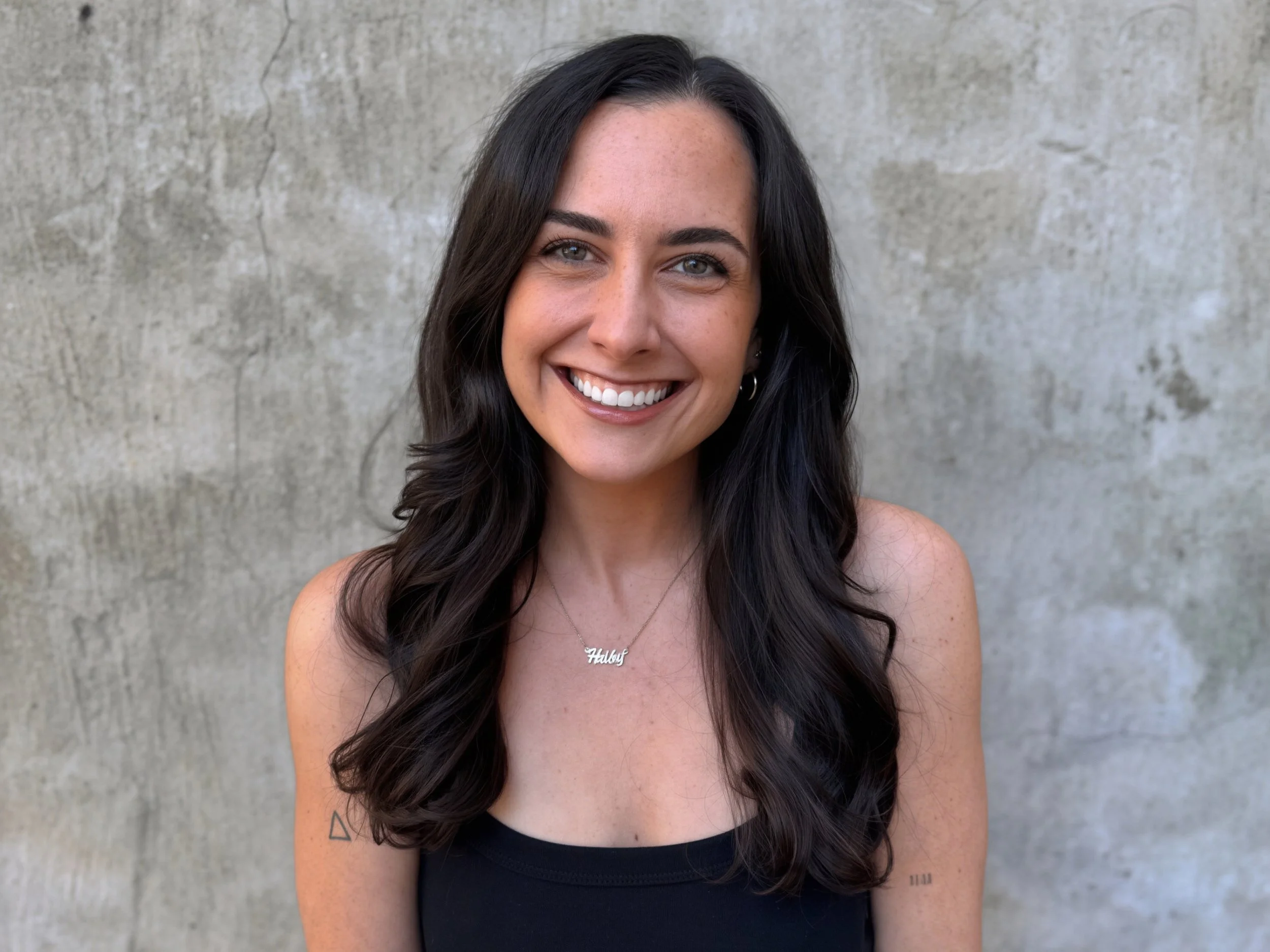 A woman with long, wavy dark hair smiling and showing her teeth, wearing a necklace with the word 'Hailey' and a black top, standing against a concrete wall.