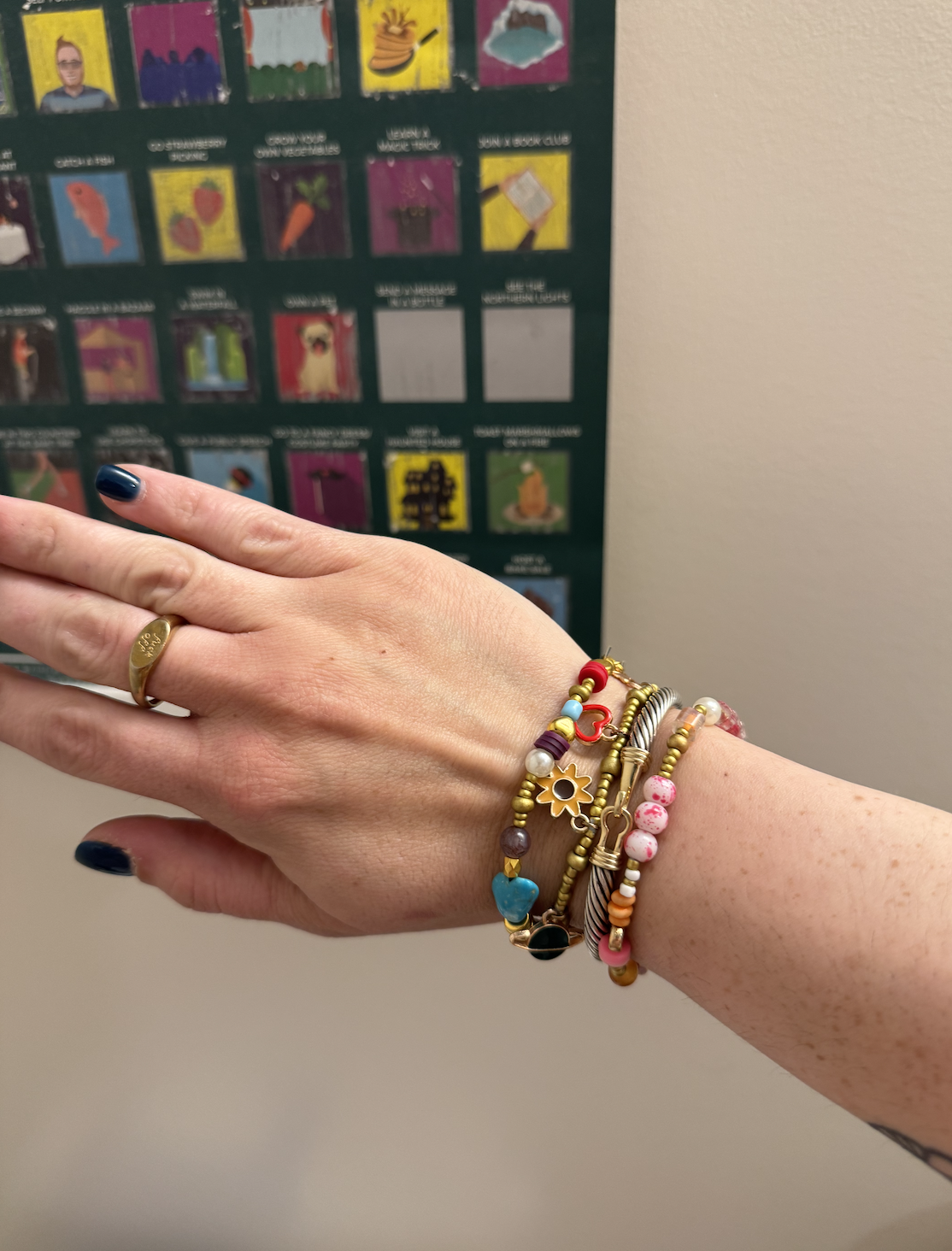 A person's left hand with navy blue nail polish, wearing a gold ring with the word 'Soul' engraved, shows multiple colorful beaded bracelets. The background has a green poster board with various colorful icons and illustrations.