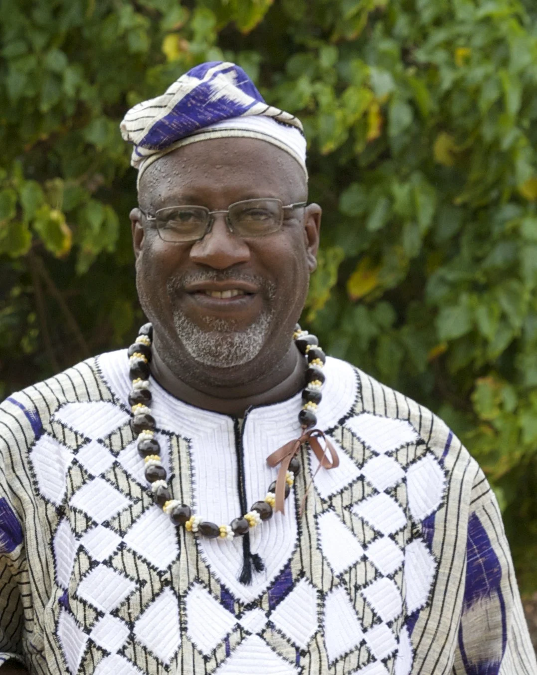 A man wearing traditional African attire, glasses, and a beaded necklace, smiling in front of green foliage.