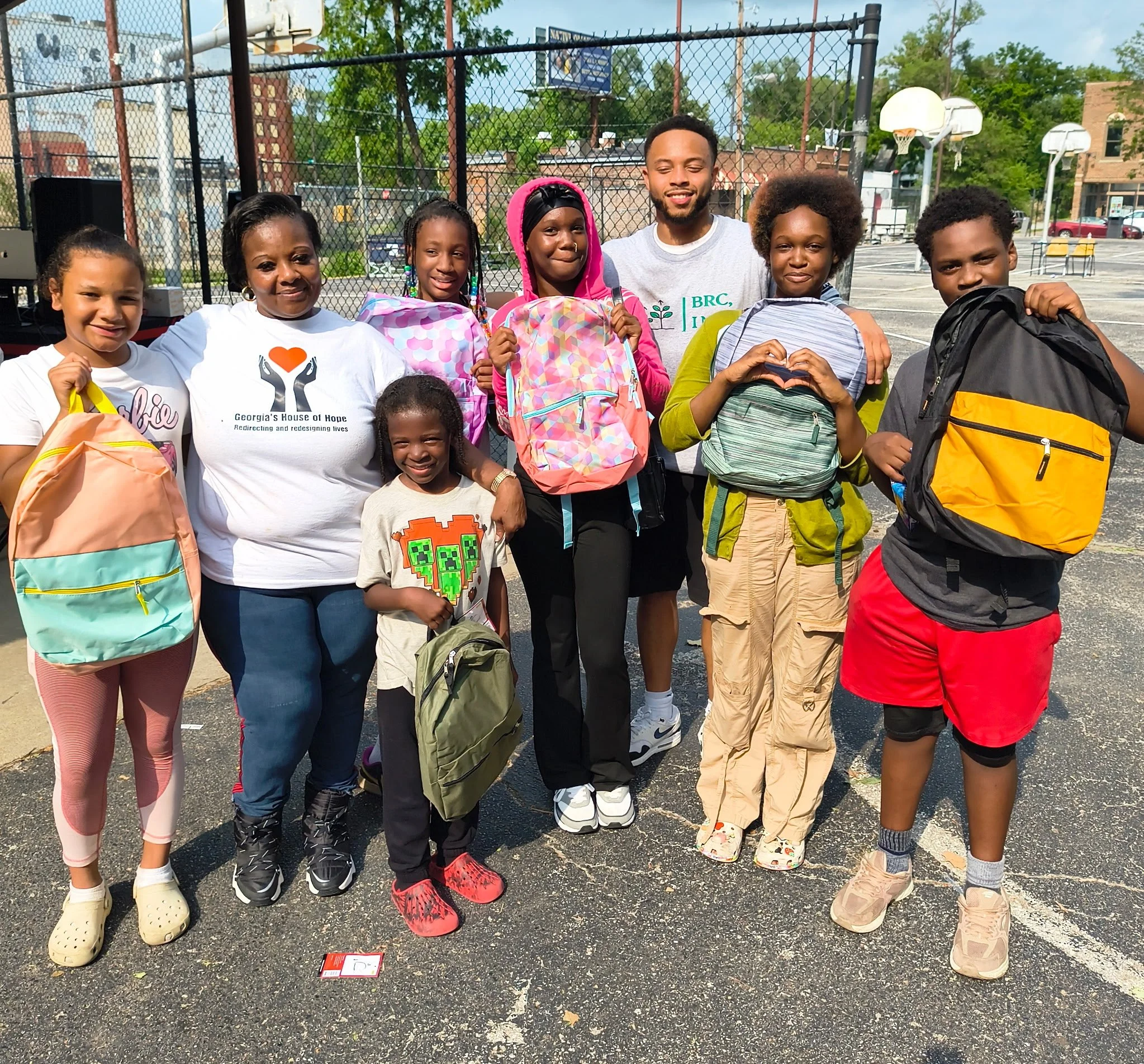 A group of children and one adult standing on a basketball court holding backpacks, smiling at the camera.