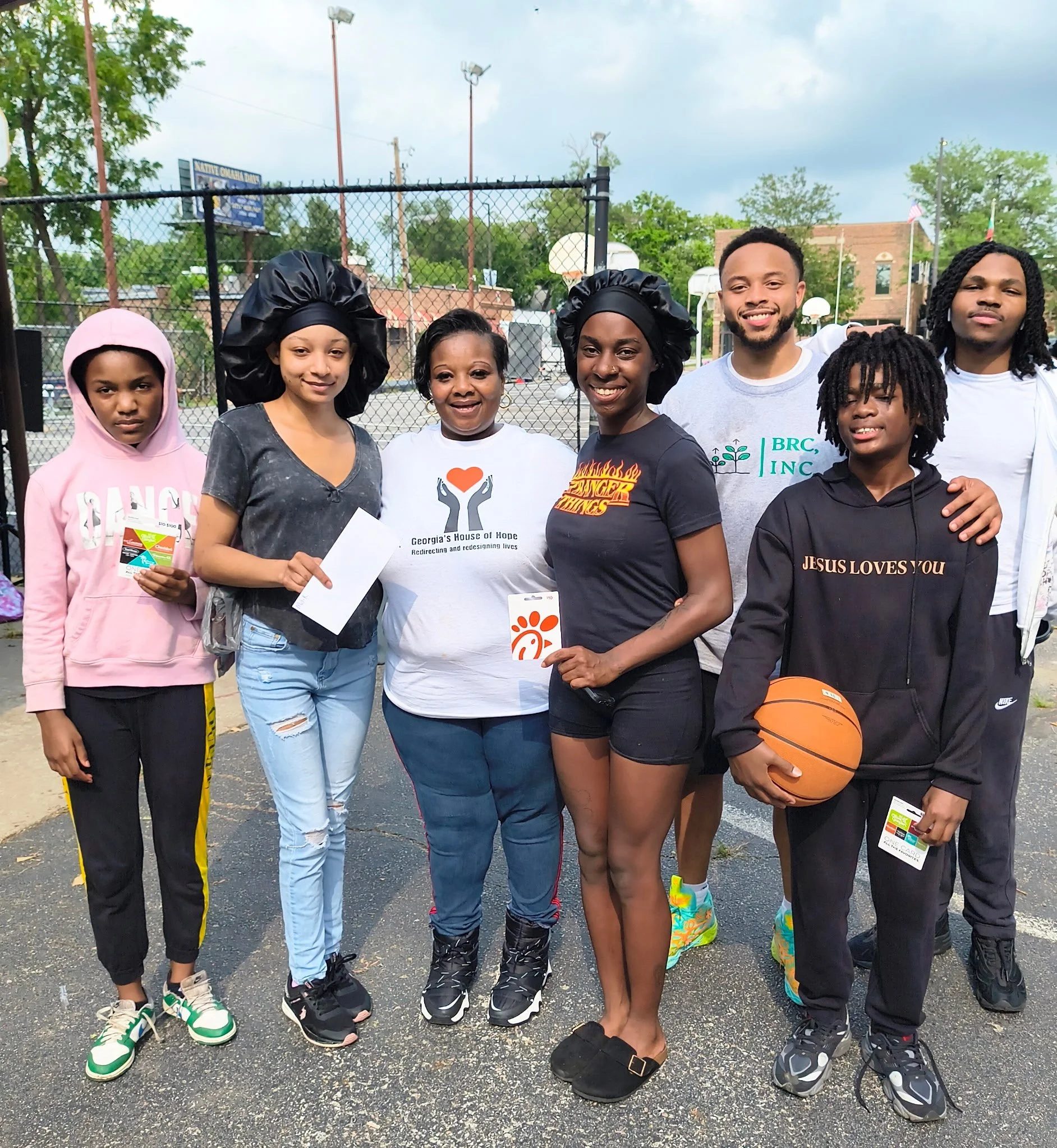 Group of seven diverse people standing on an outdoor basketball court, smiling at the camera, with some holding a basketball and pamphlets.