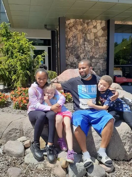 A man and three children sitting on a large rock outside a building with stone and glass walls. The man is smiling and has his arms around two children, while another girl sits close to him. All are casually dressed.