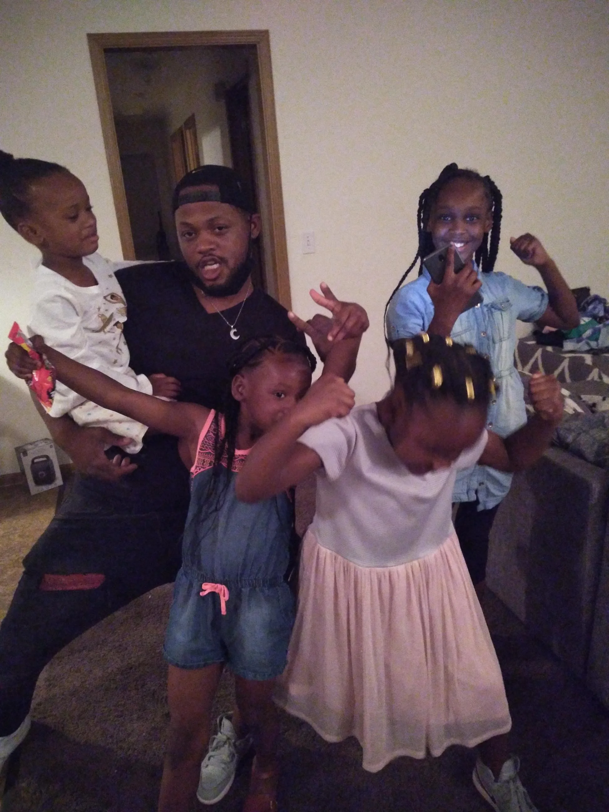 A man and four young girls posing together in a living room, making playful gestures and smiling.